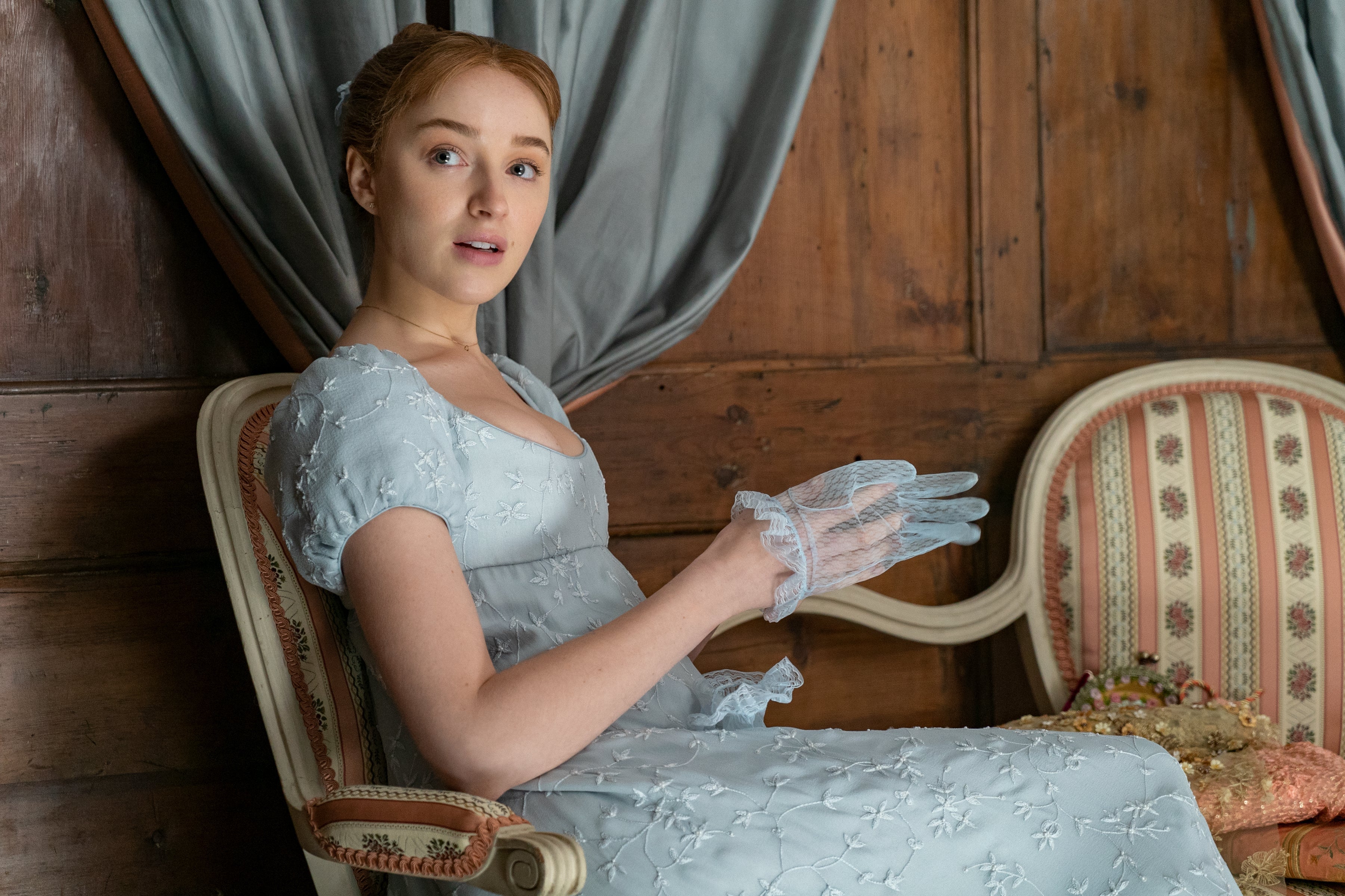 Phoebe Dynevor sits in a chair glancing over her shoulder. One hand is halfway in a translucent blue glove.