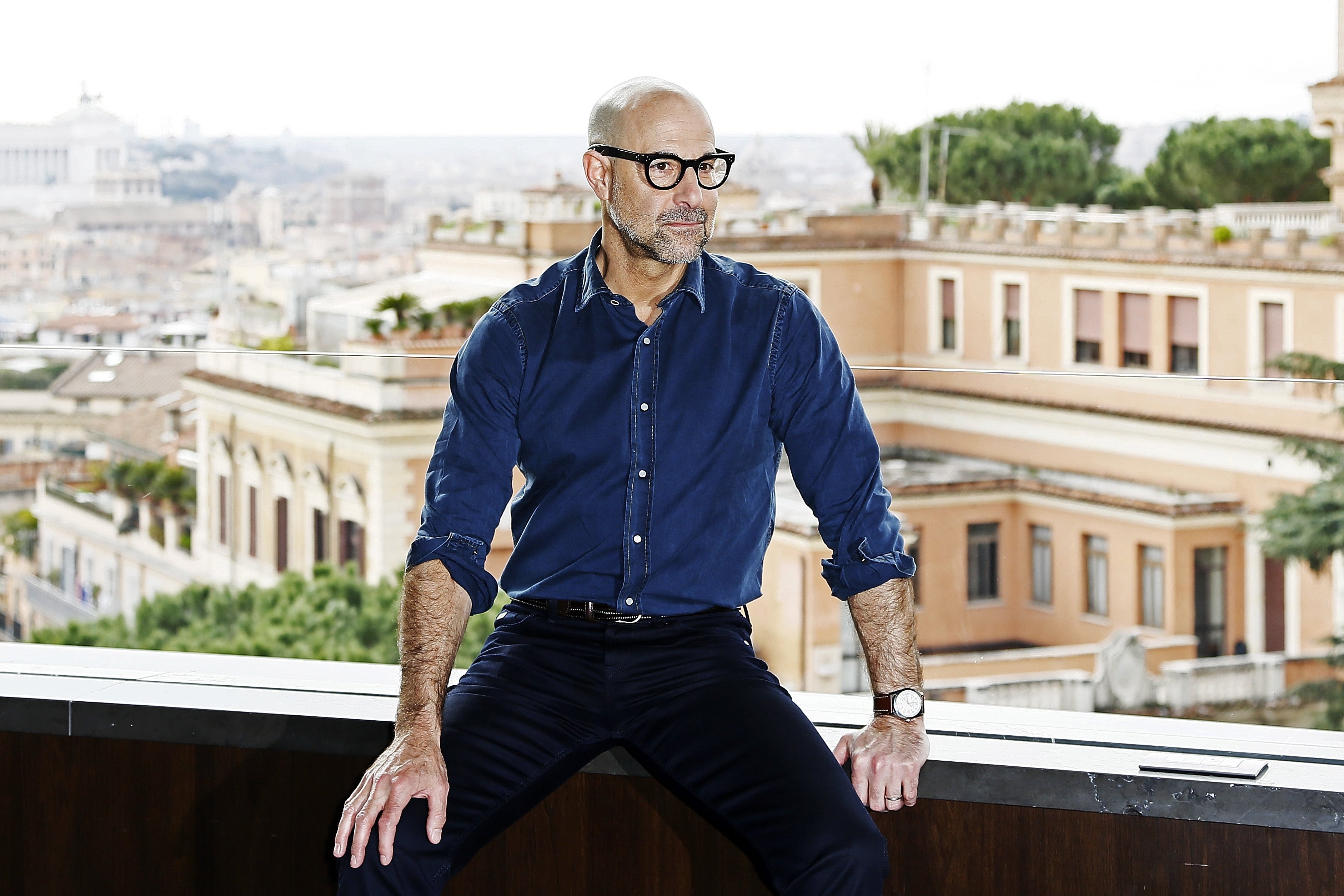 Stanley Tucci sitting on a ledge overlooking Italian buildings, his sleeves rolled up to reveal his famous forearms.