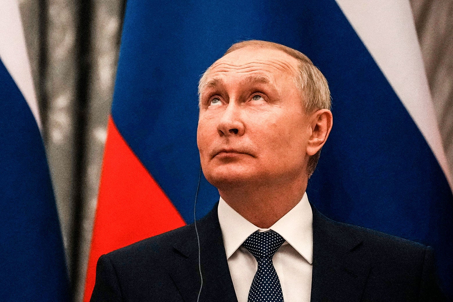 Vladimir Putin looks upward while standing in front of a Russian flag.