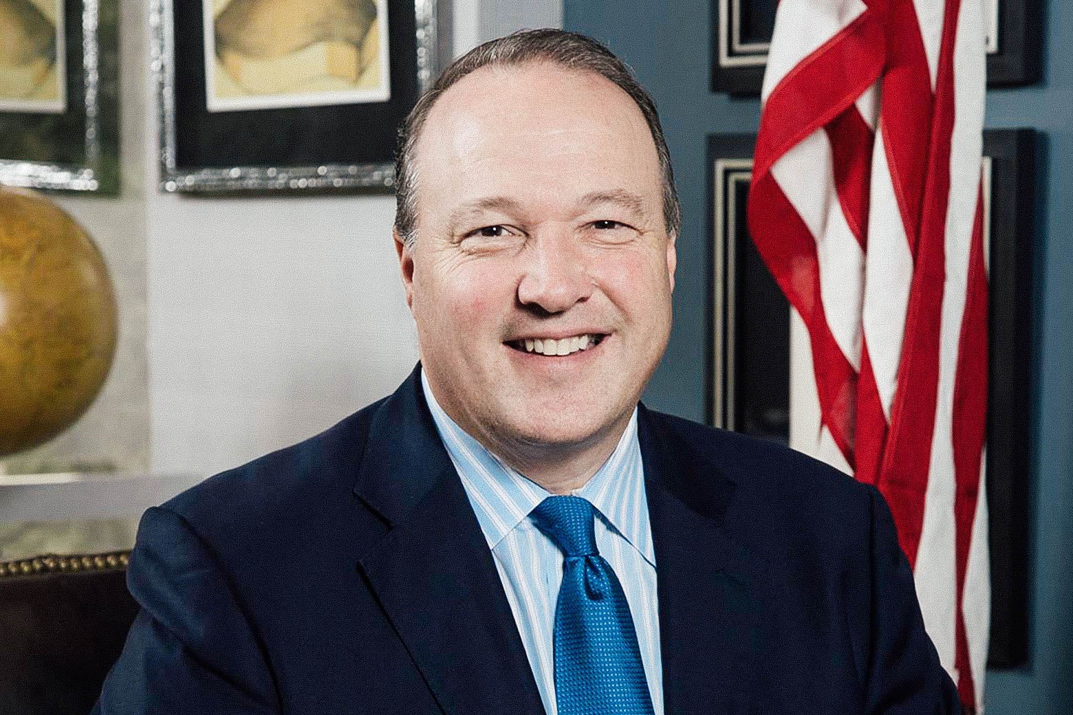 A smiling besuited man in an office with an U.S. flag 