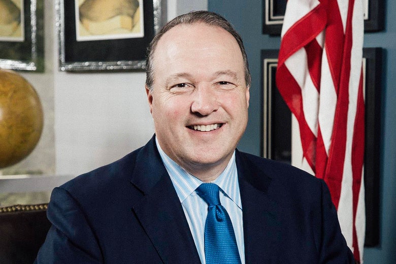 A smiling besuited man in an office with an U.S. flag 