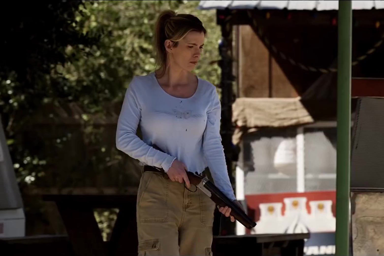 Betty Gilpin standing in front of a country gas station, holding a shotgun.