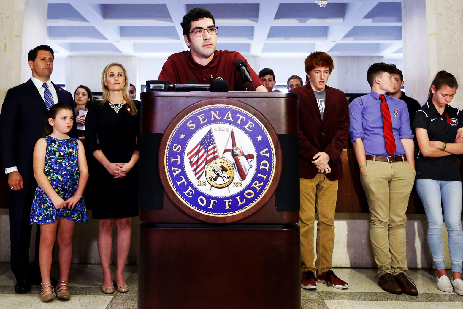 Lorenzo Prado stands at a podium with fellow survivors and family members behind him.