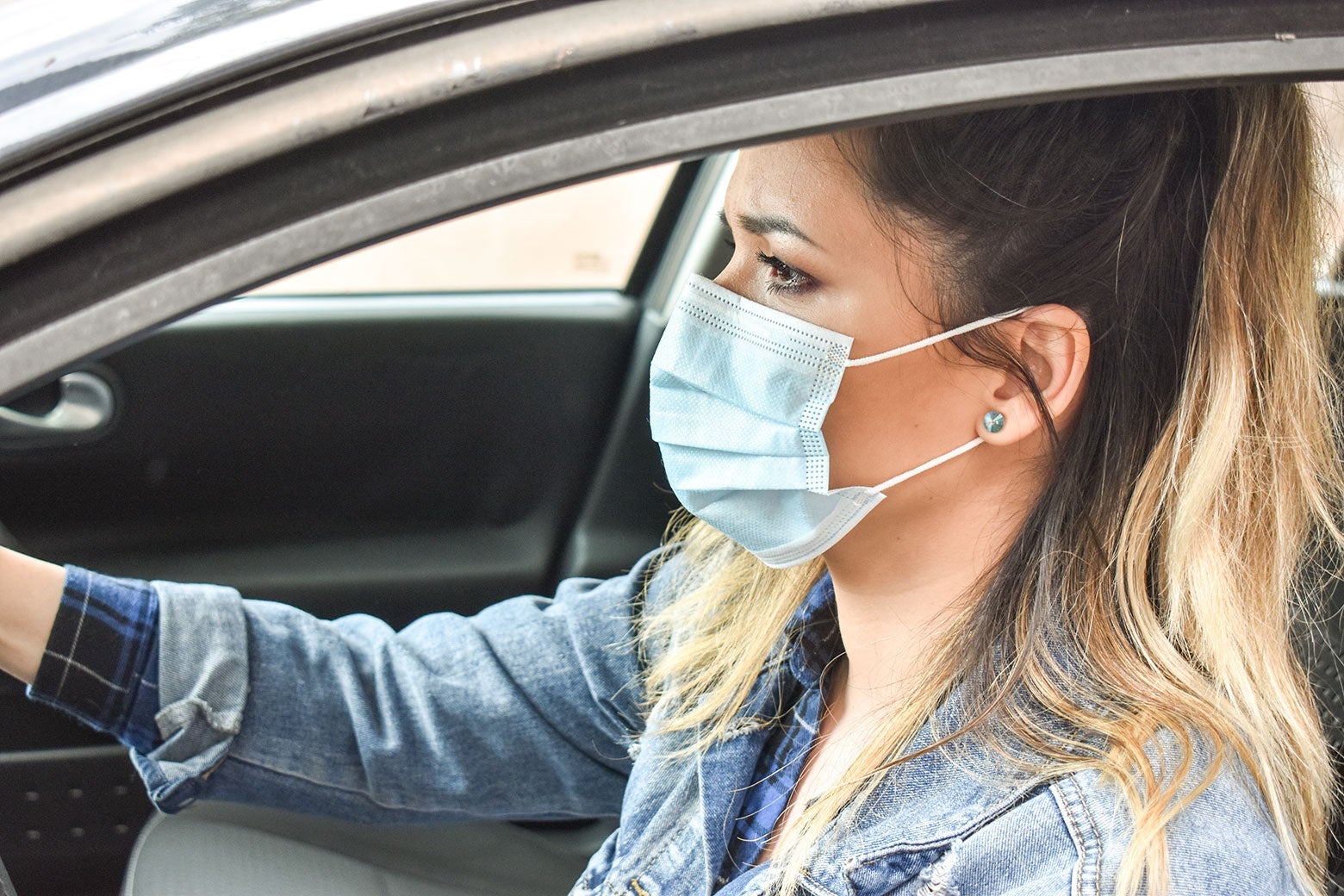A woman wearing a surgical mask, sitting at the wheel of a car