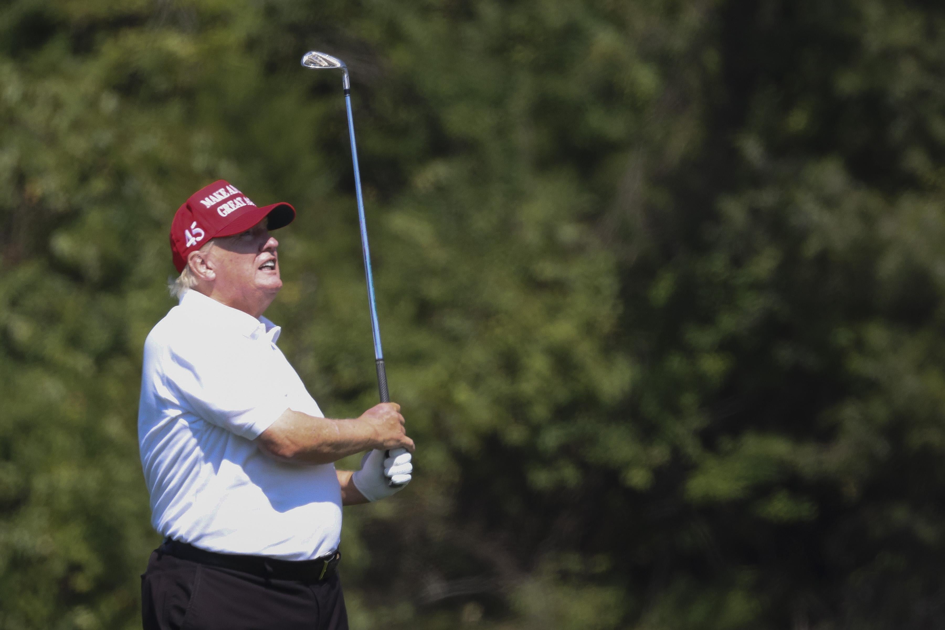 Trump, in a white polo and a red ball cap, holds a golf club aloft as he watches his shot. 
