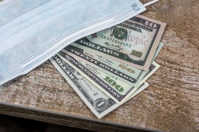 A light blue surgical masks covers a 10, 20, 100, and 1 dollar bill fanned out on a wooden table.