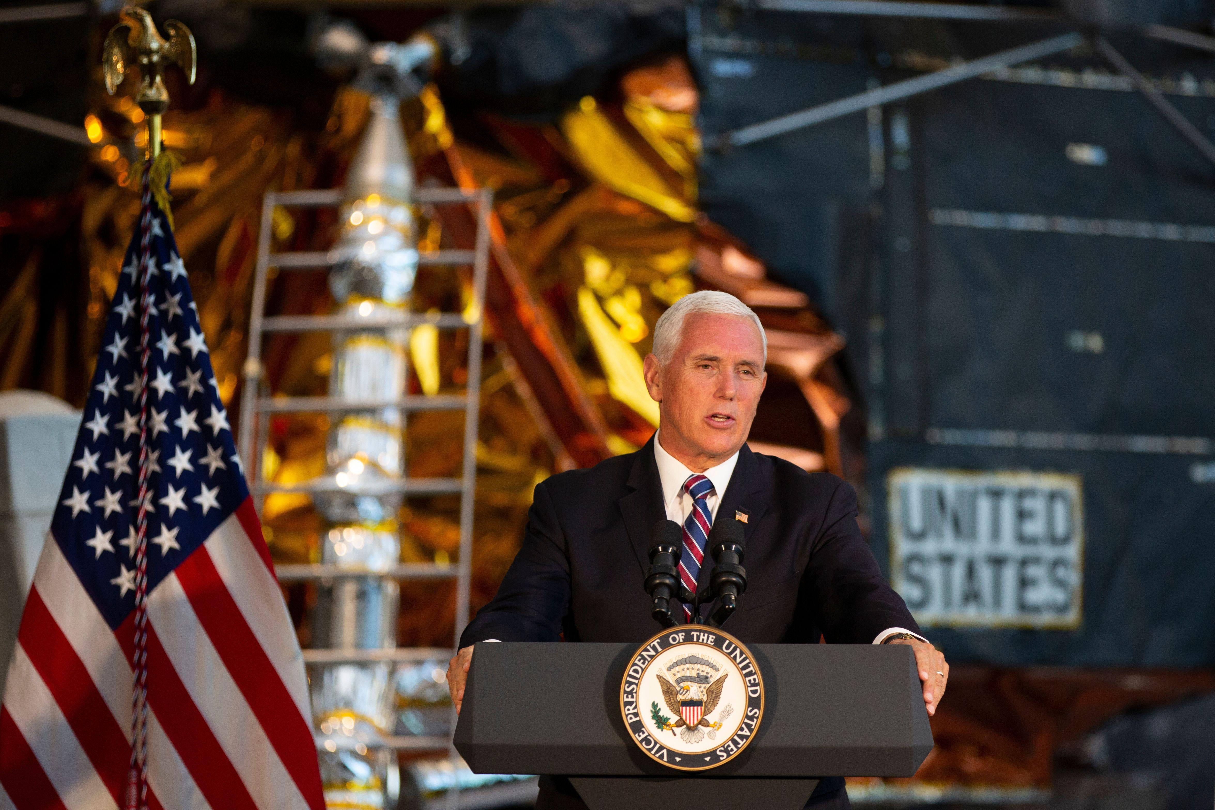 Pence stands before a podium, with the American flag and spacecraft in the background.