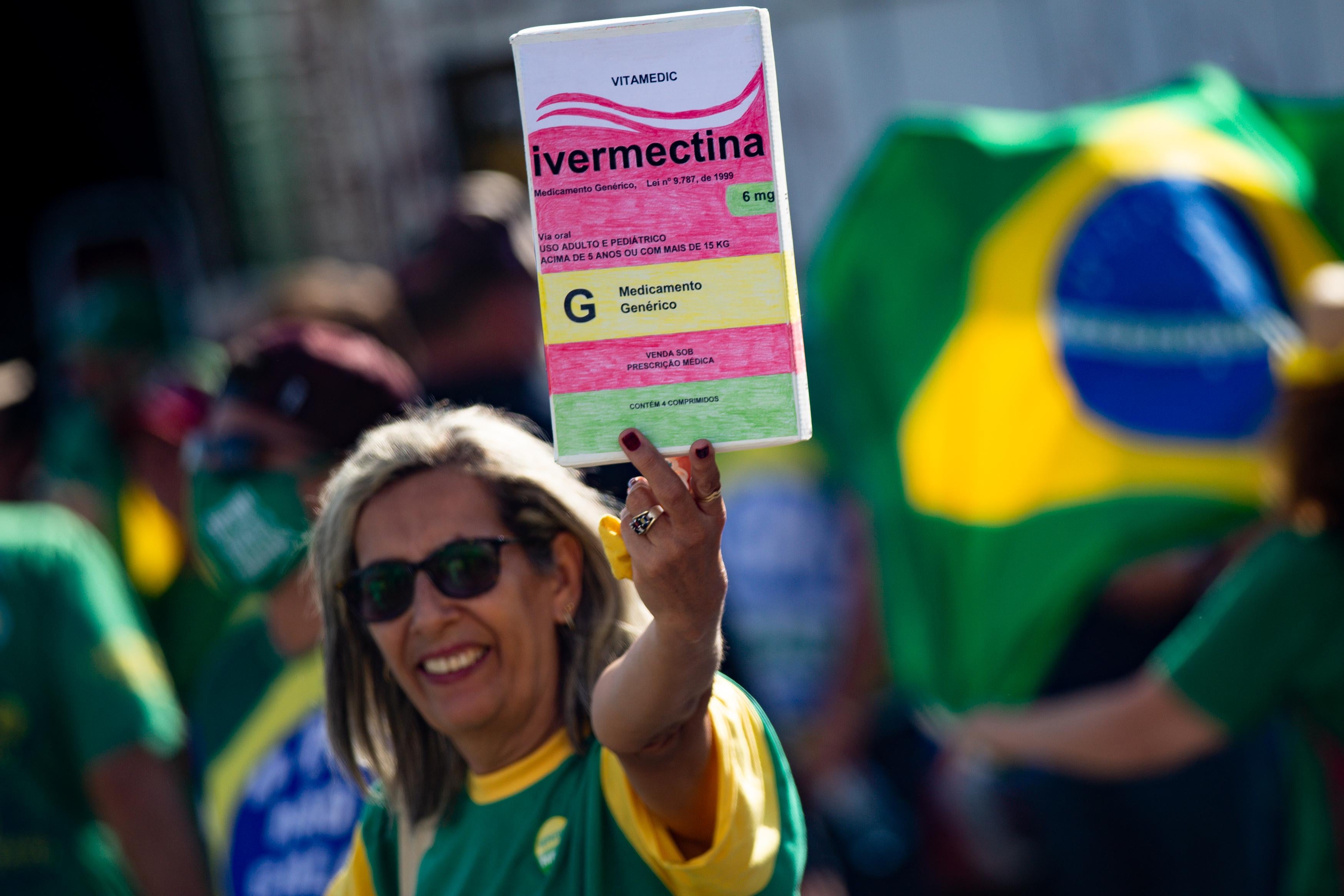 A woman holds a large box of box of ivermectin while someone behind her unfurls a Brazilian flag.