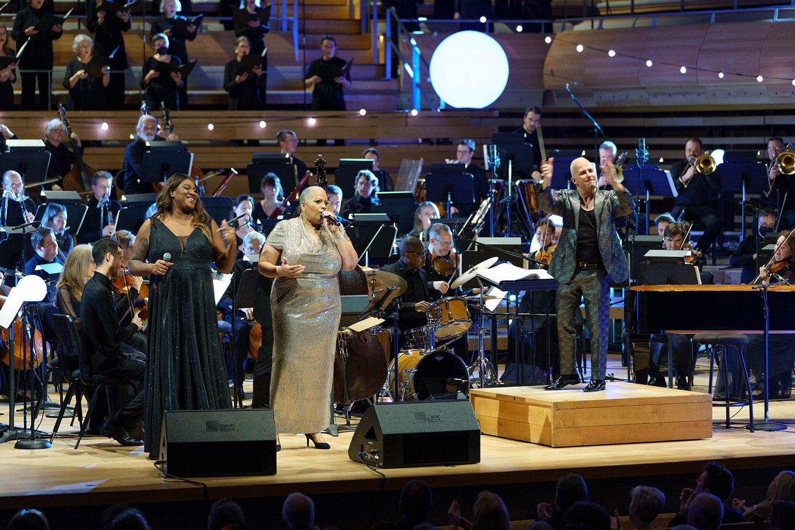 Smiling singers in formalwear and an orchestra, conductor, and white holiday string lights onstage at Canada's Maison Symphonique.