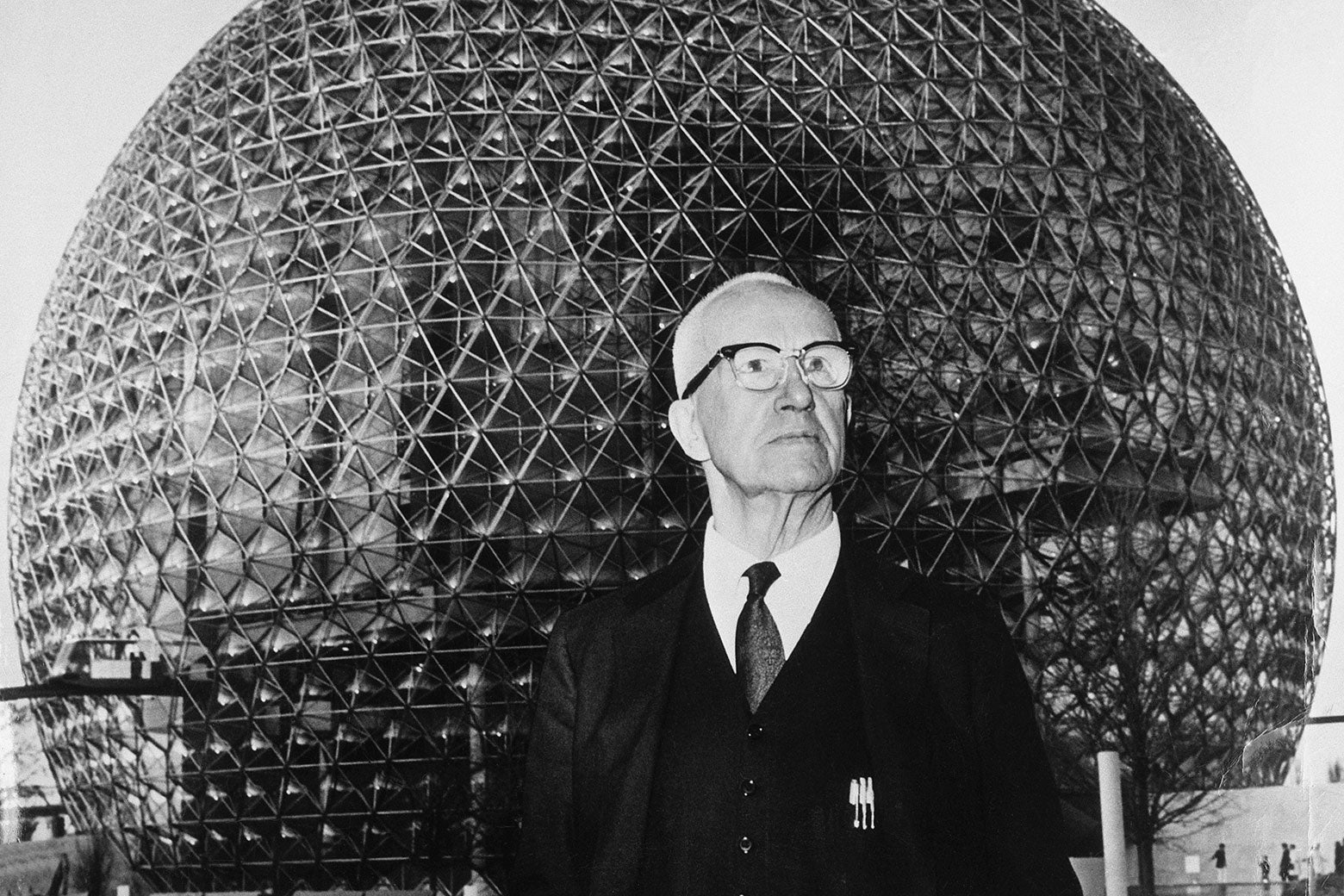 Buckminster Fuller dressed in a suit and tie and wearing glasses gazes into the distance in this black and white photo of him in front of a geodesic dome at the 1967 World's Fair.