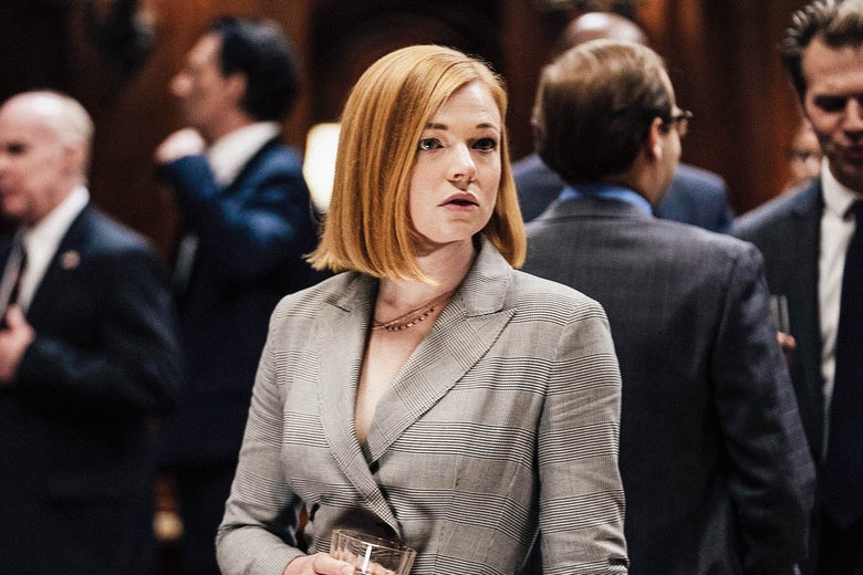 A redheaded woman stands in the middle of a crowd holding a drink and looking to the side.