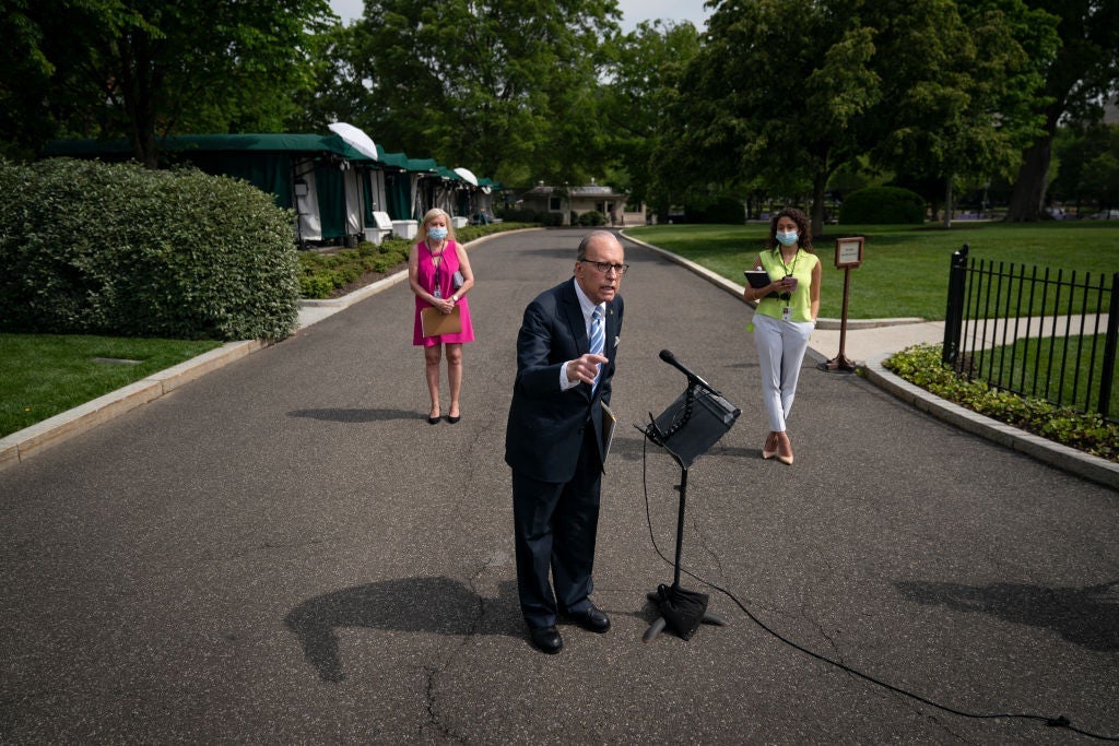 Kudlow leans over to speak into a microphone set up in the middle of a road as two women wearing masks stand at a distance behind him.