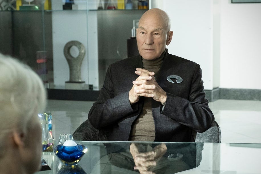 Patrick Stewart sits with his hands folded at a desk across from a white-haired woman seen from behind. 