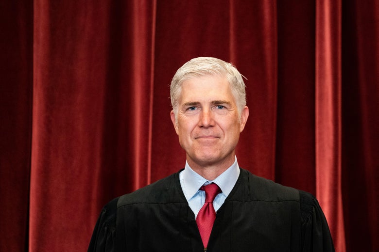Neil Gorsuch stands smiling in his robes in front of a red velvet curtain.