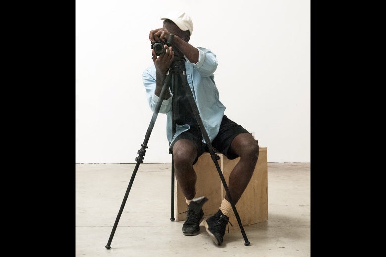A casually dressed young black man is taking a photograph. The camera is on a tripod.
