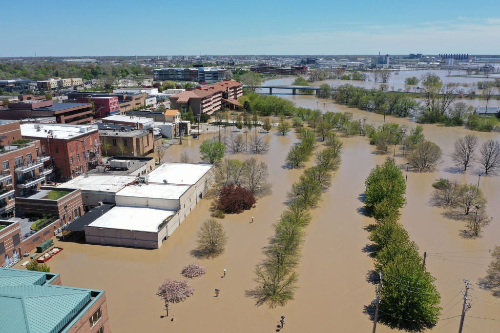 An aerial shot depicts brown river water that has flooded up to the edge of a small-town downtown, submerging buildings and trees.