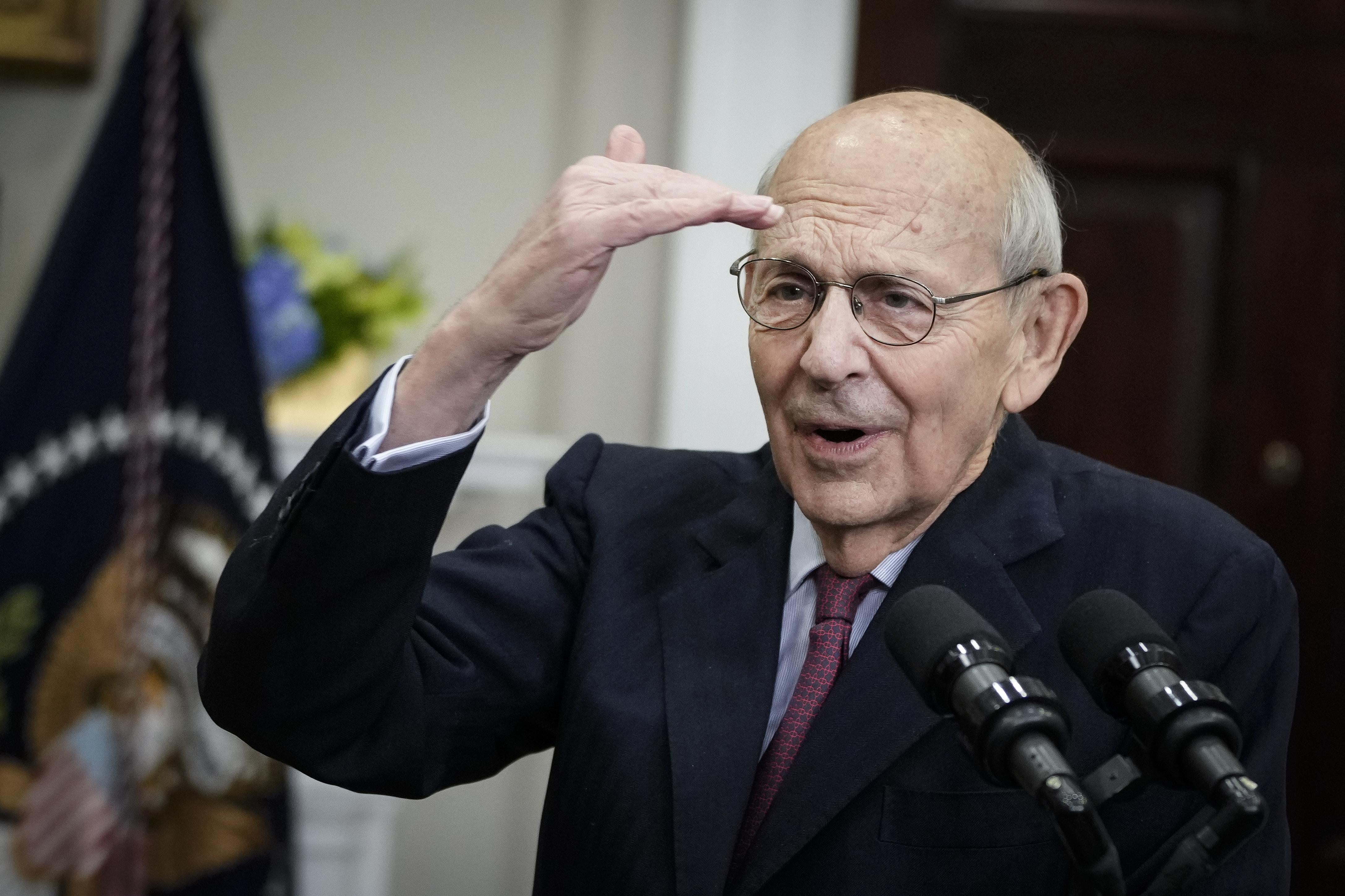 Breyer in a suit gestures towards his head with his right hand.
