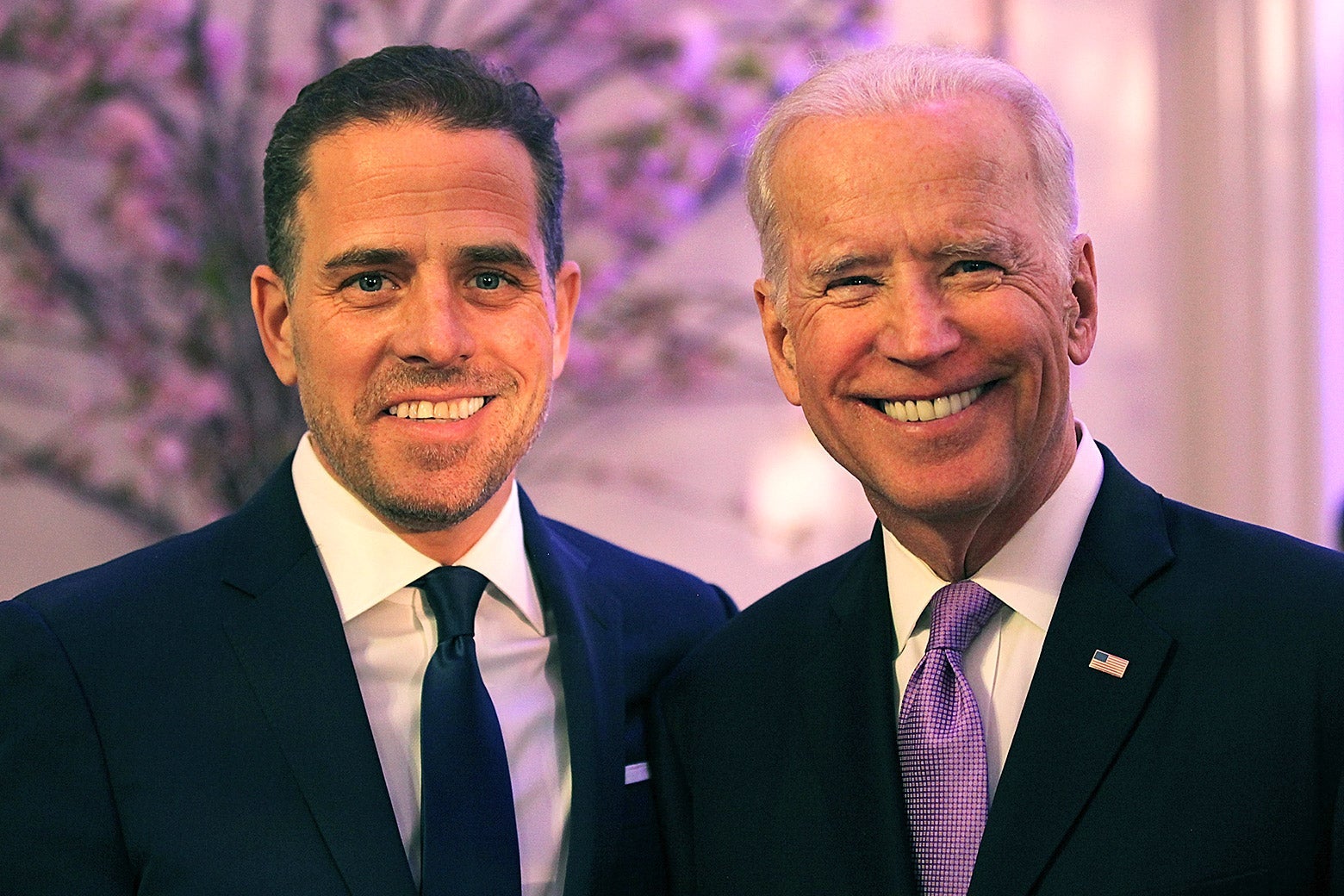 Hunter and Joe Biden smile toward the camera, standing in a room with light purple lighting and a tree in bloom in the background