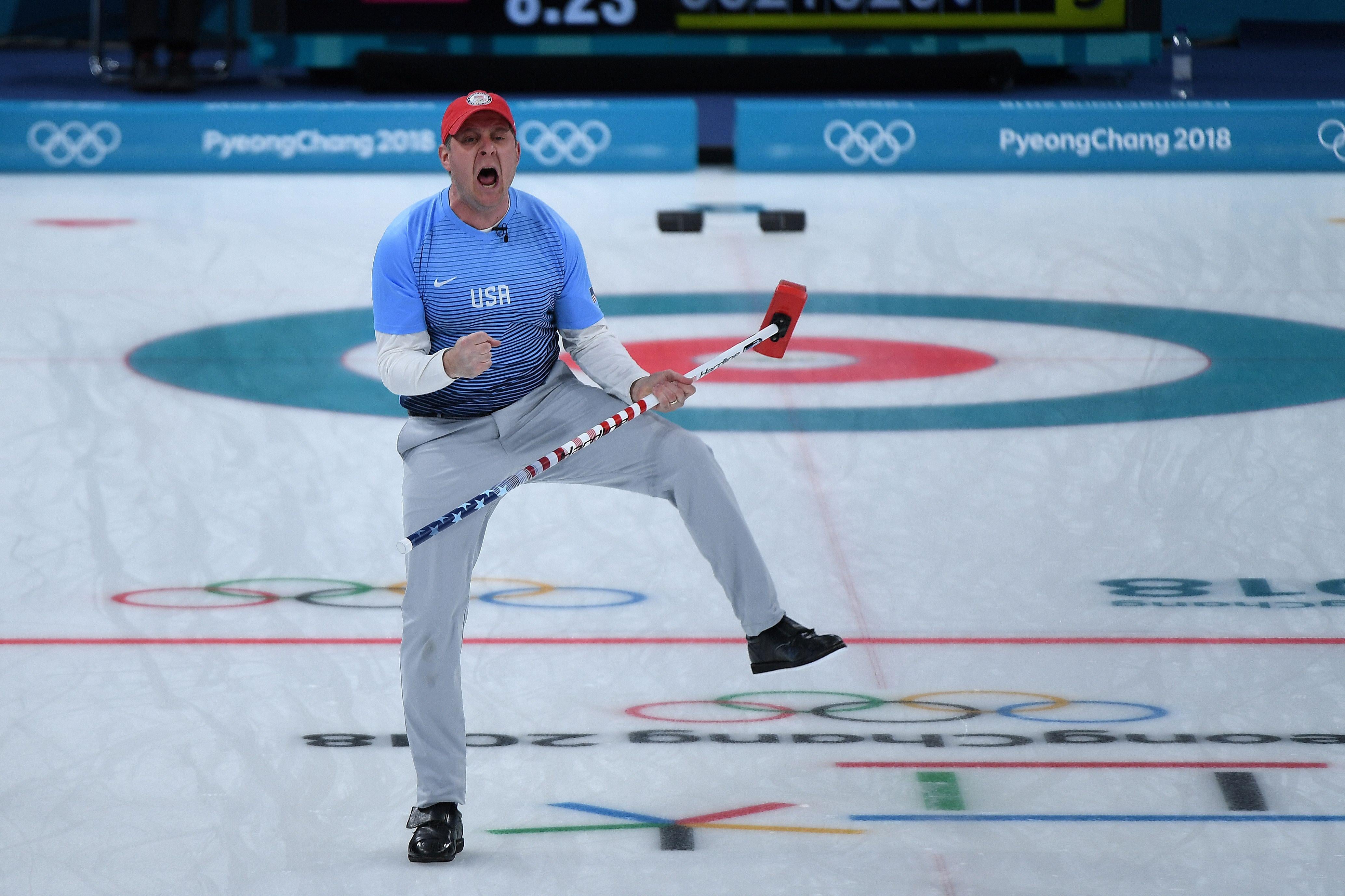 John Shuster, holding his curling broom, stands on one leg and yells in triumph.