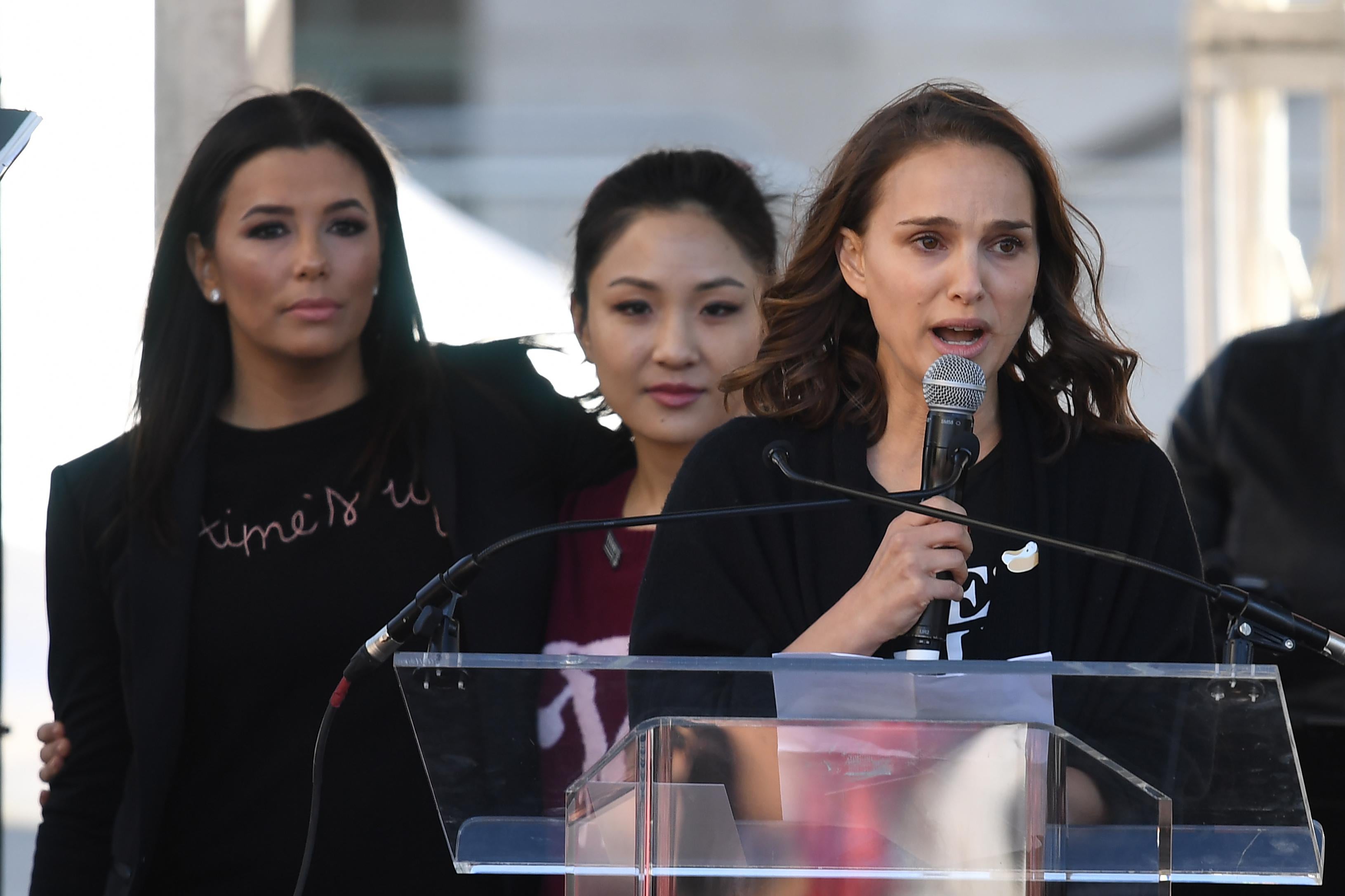 Natalie Portman speaks at a podium while actors Eva Longoria and Constance Wu stand behind her.