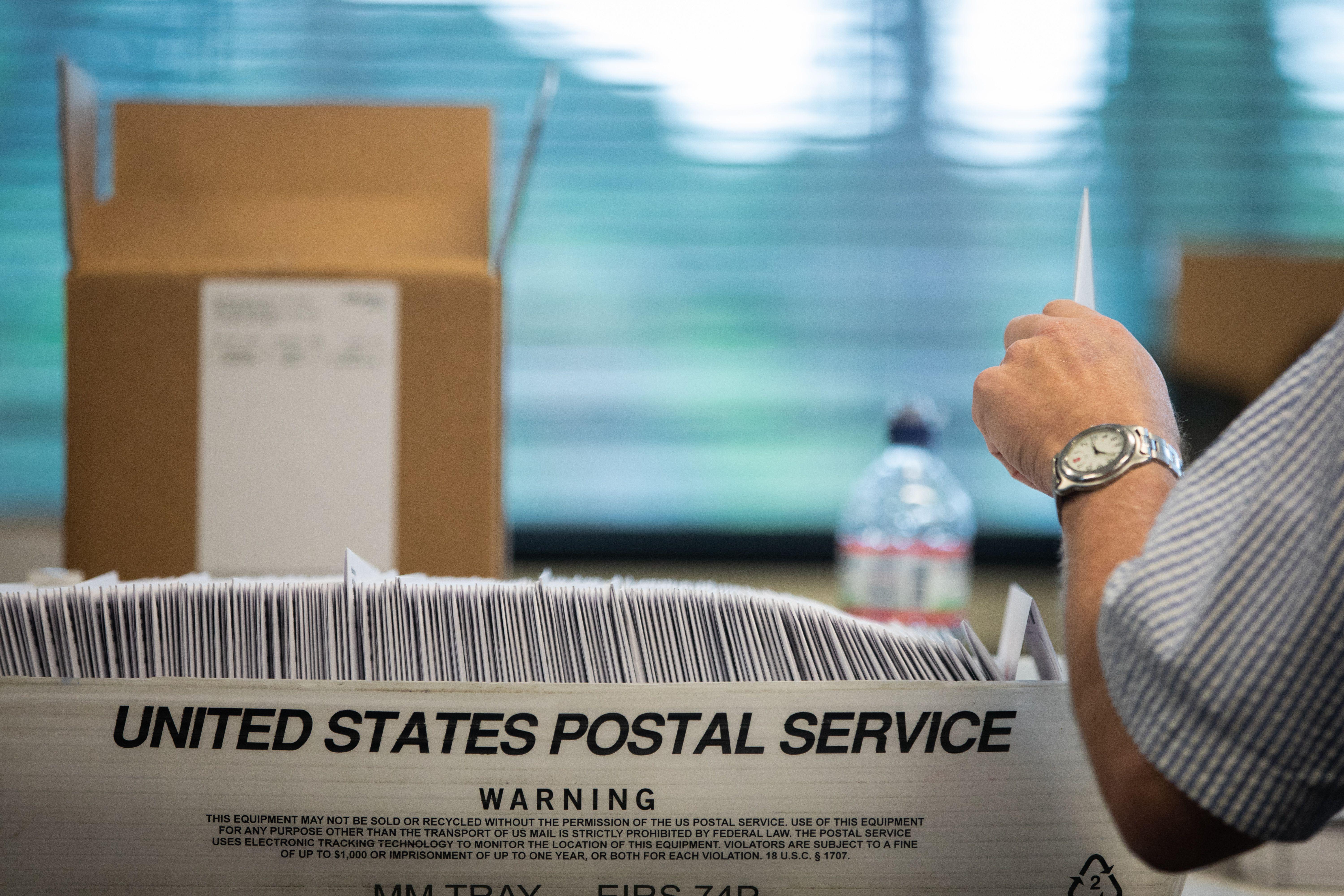 A box reading "United States Postal Service" is seen full of files. An arm is seen taking one out.
