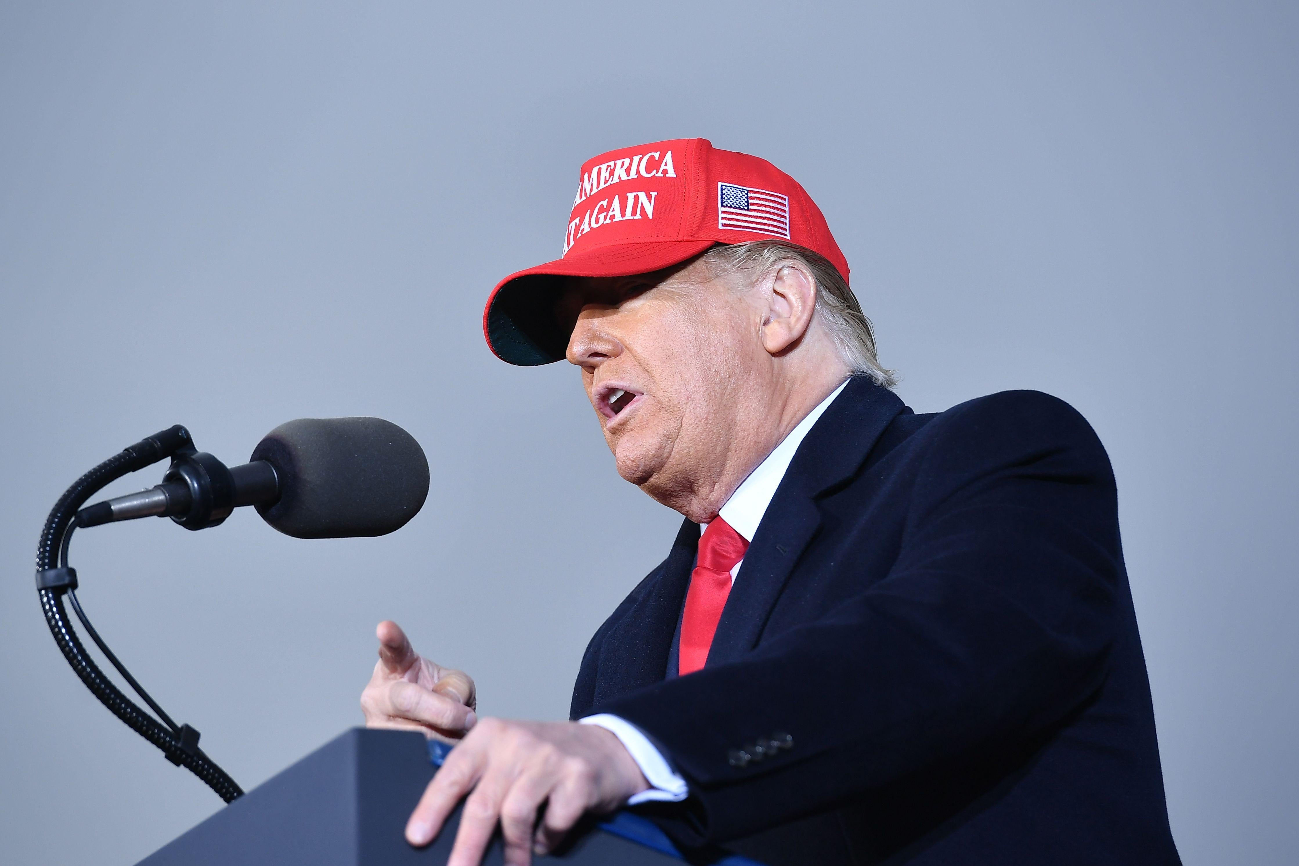 Donald Trump speaks into a microphone at a podium while wearing a red MAGA hat.