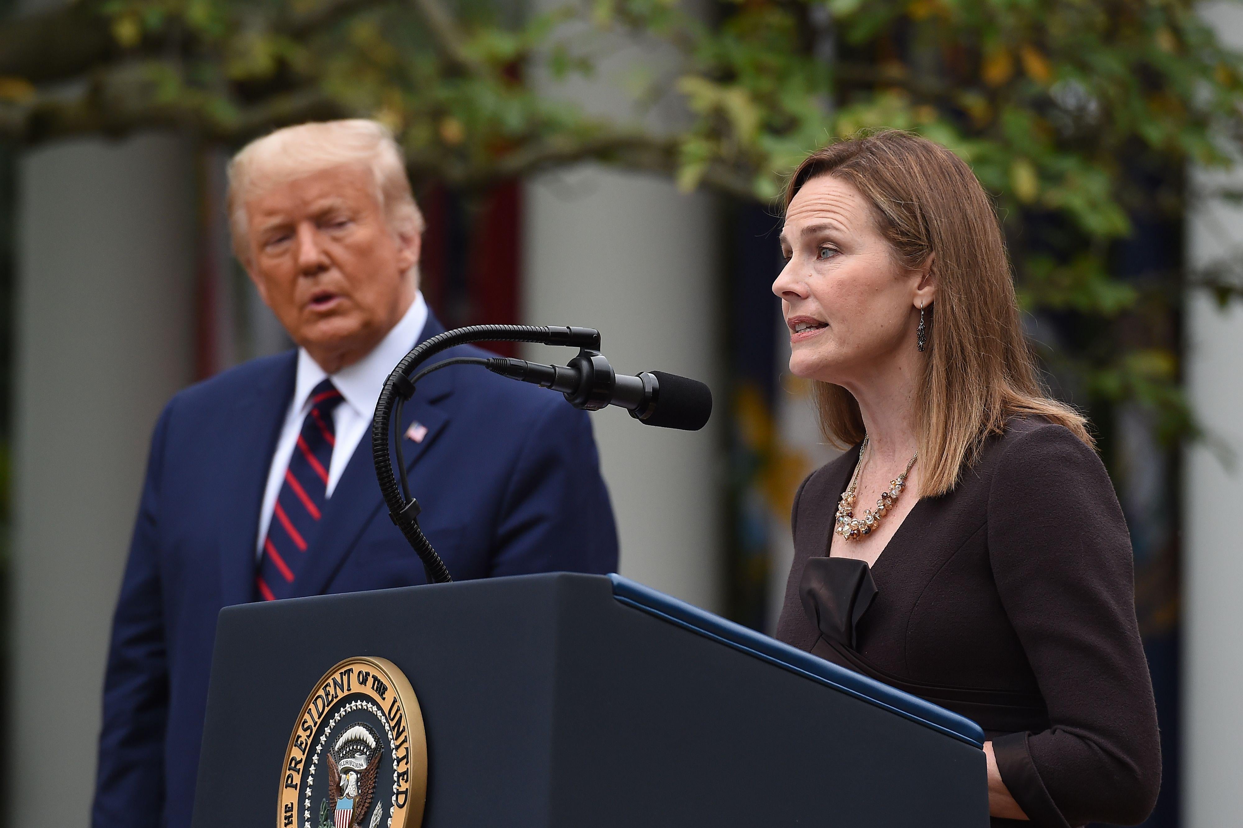 Amy Coney Barrett speaks at a lectern while Trump watches from the side.