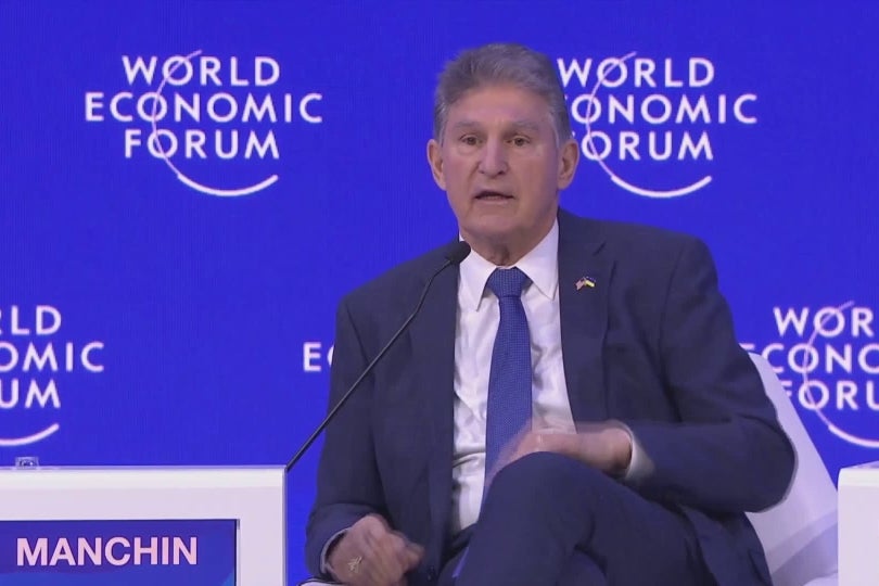 Manchin speaks into a long skinny mic, seated and cross-legged, with the World Economic Forum logo on a screen behind him