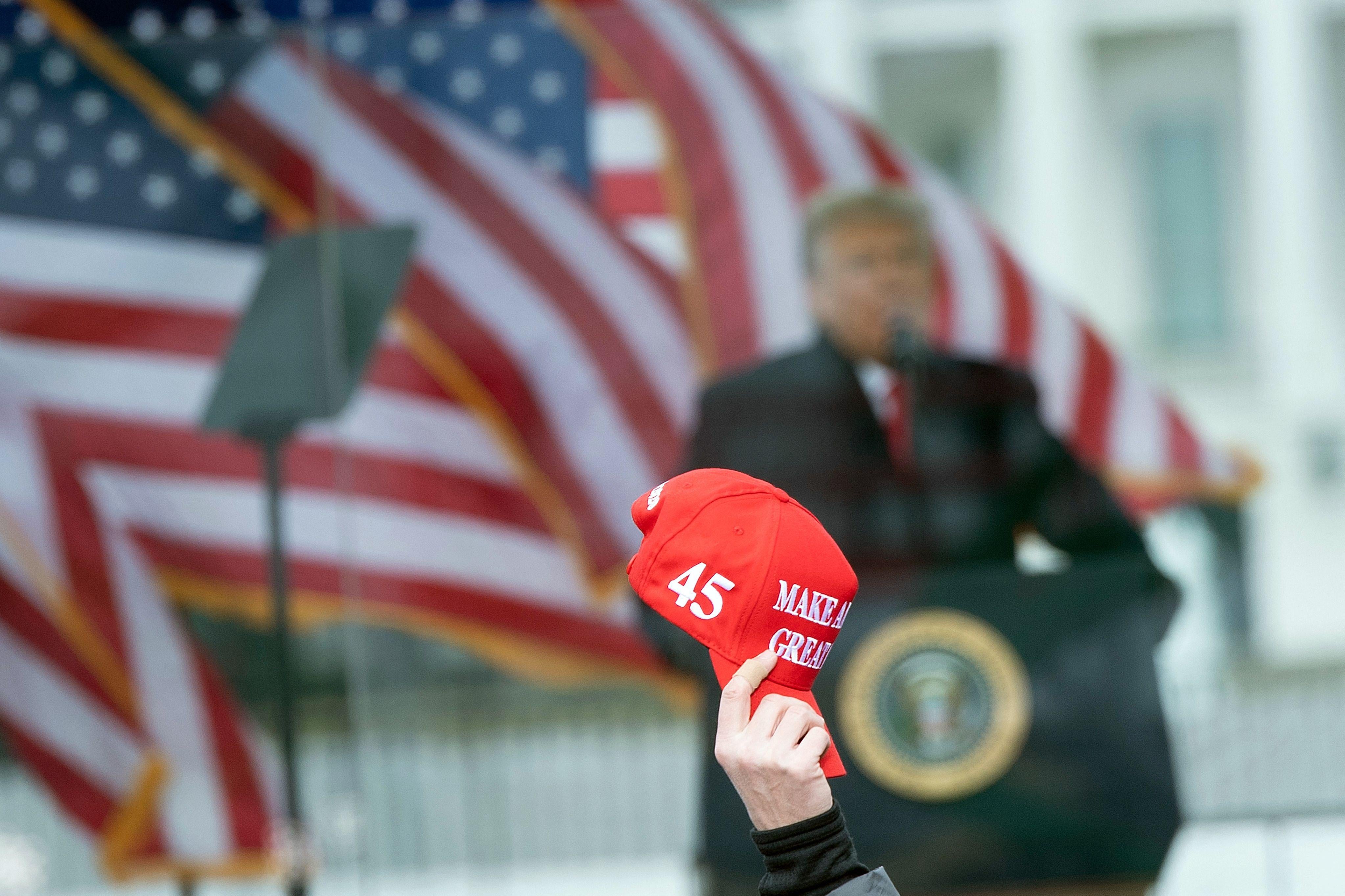 Trump standing at a lectern beside American flags out of focus in the background and a hand holding up a red MAGA hat in focus in the foreground