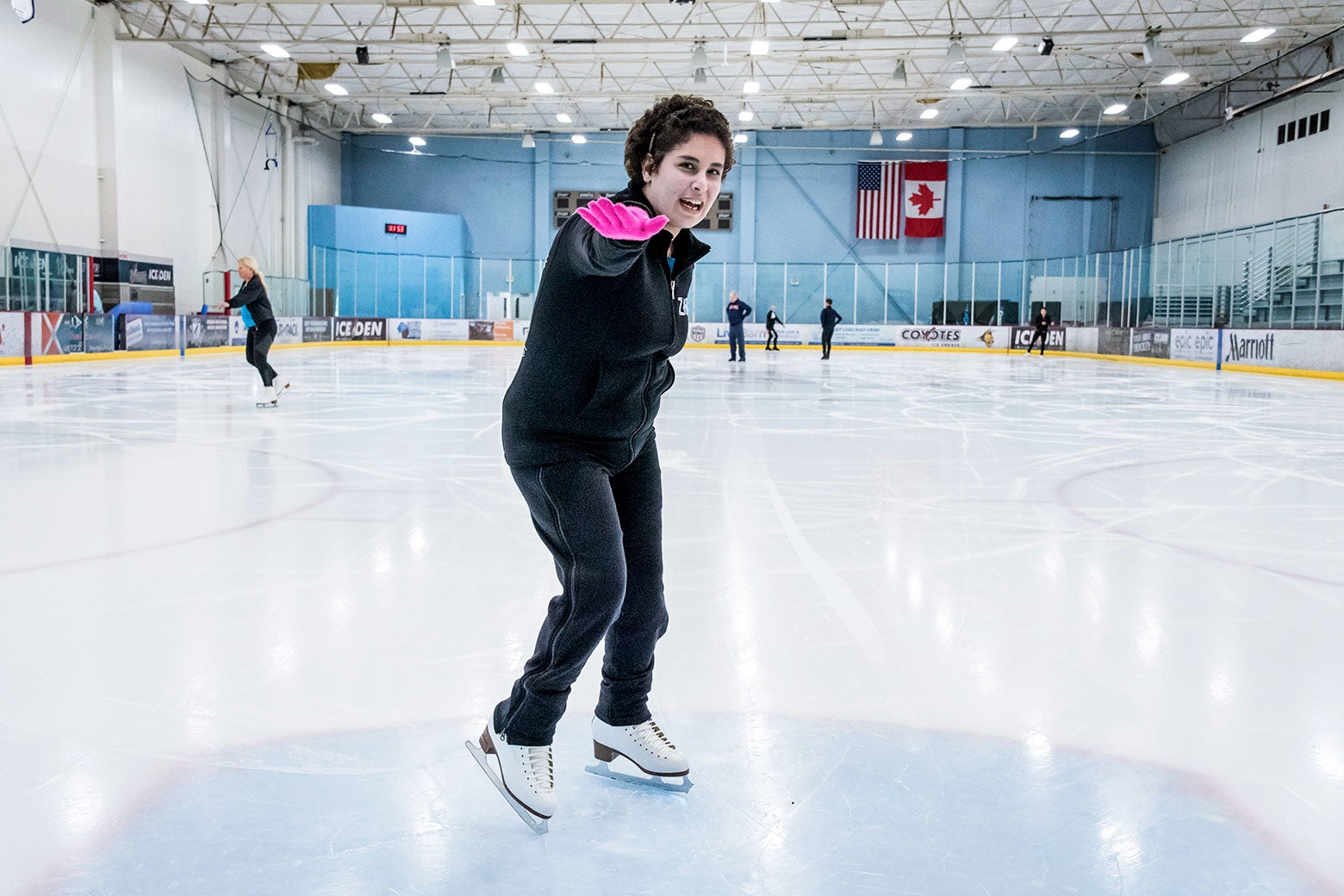 A woman on ice skates reaches a pink-gloved hand out toward the camera as she smiles.