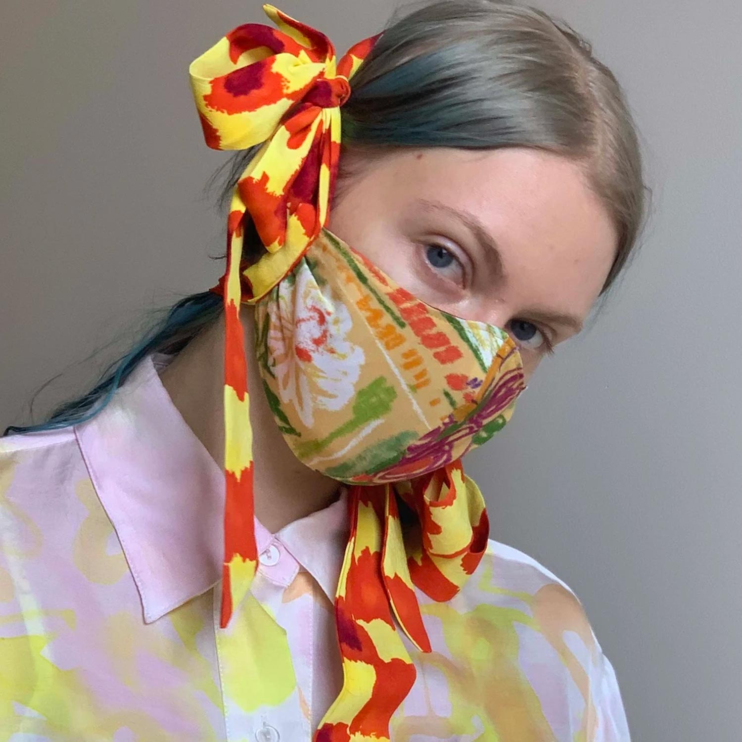 A model with blue hair wears a colorfully patterned face mask with large bows that loop around her head.
