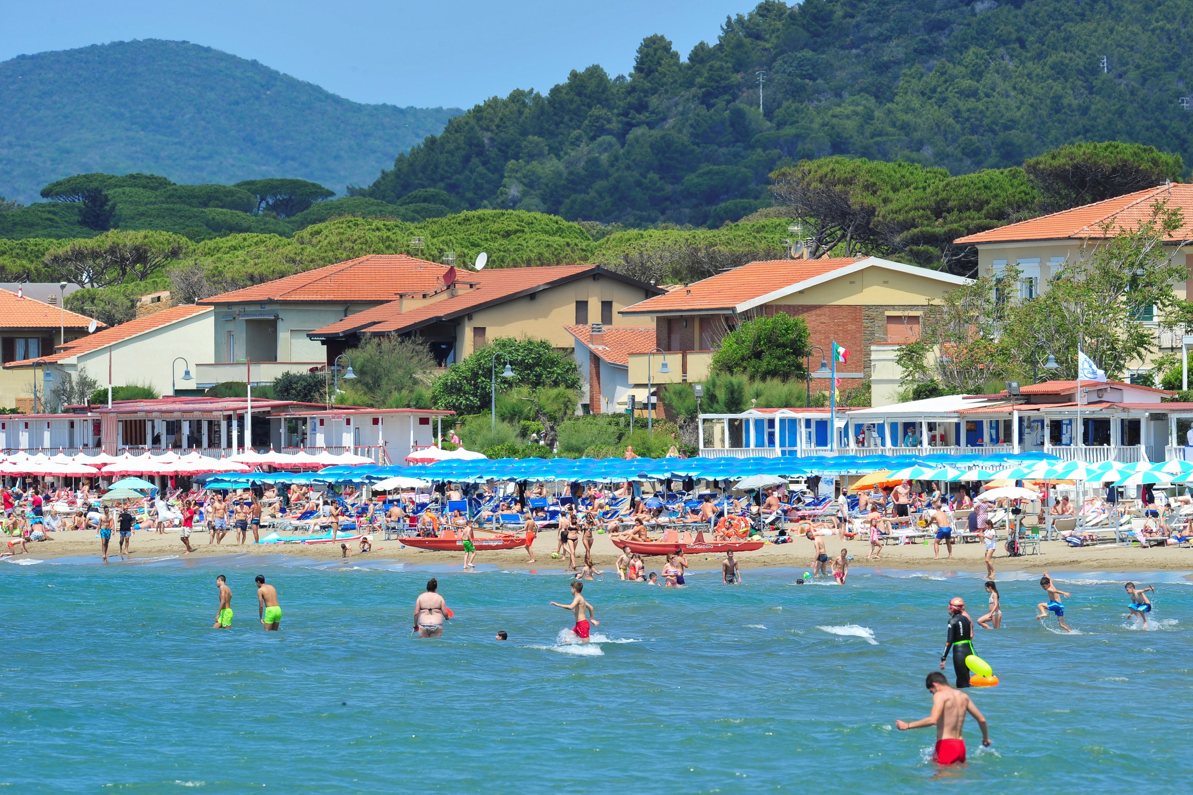 A view of the shore from the sea, with swimmers, sunbathers, and small beach houses in the background.
