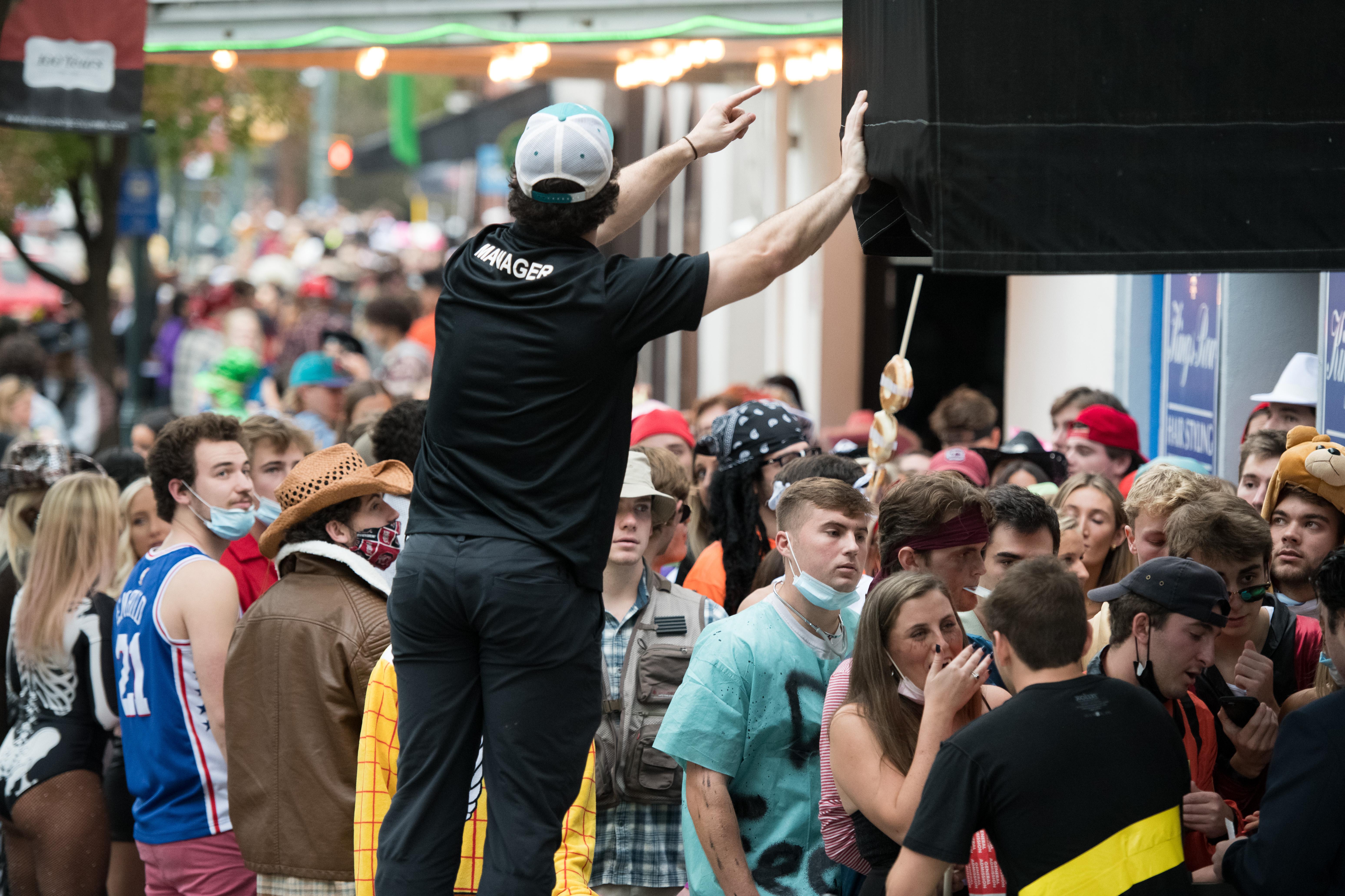 A night club worker stands on a chair above a crowded group of people, most of whom are maskless or wearing their masks around their chins.