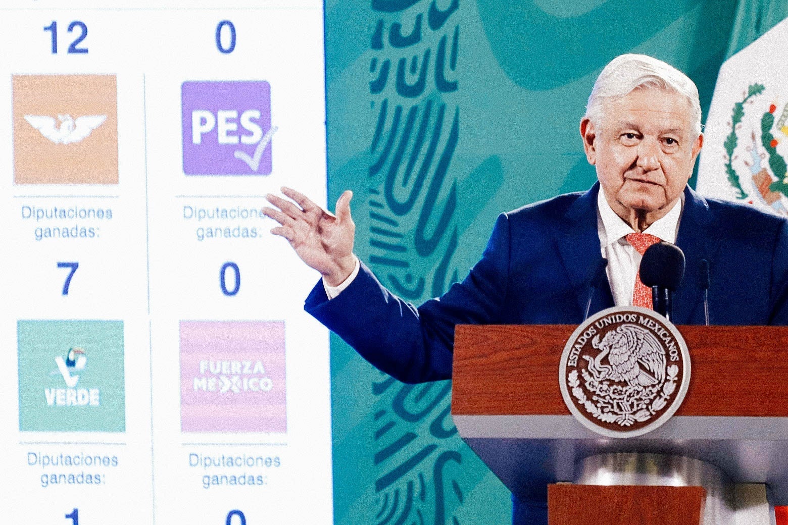Lopez Obrador stands at a podium gesturing with one arm beside a poster displaying the logos of Mexican political parties, the Green Party among them