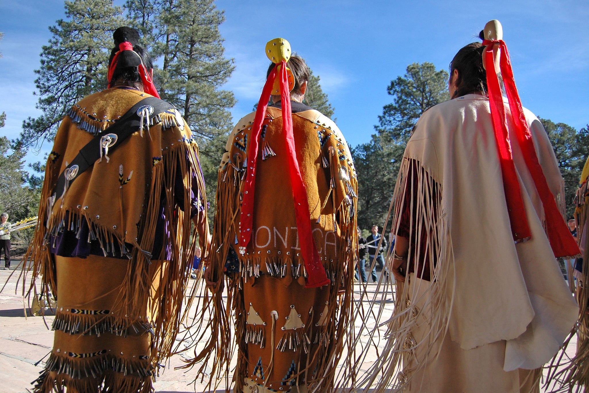 Indigenous people in ceremonial dress are seen gathered in front of pine trees.