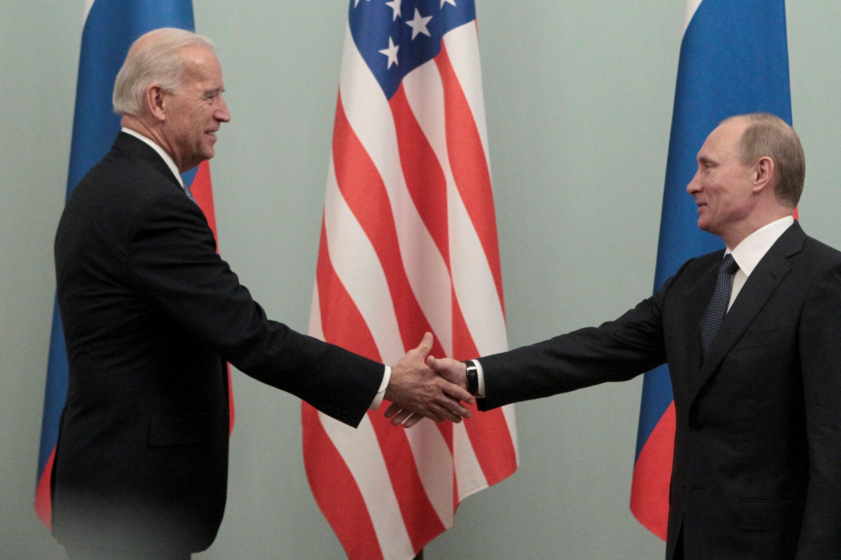Biden and Putin smile and shake hands in front of American and Russian flags.