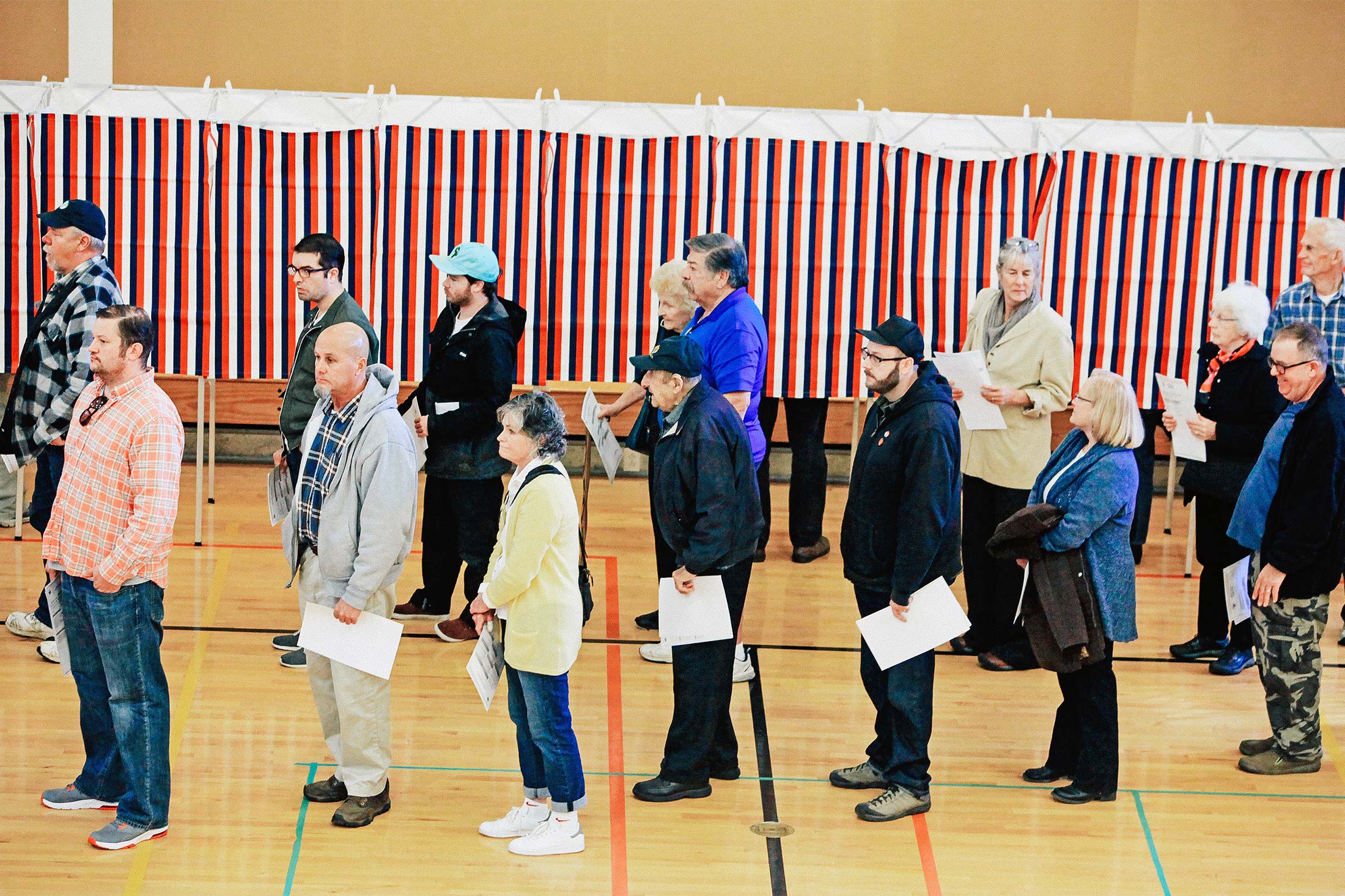 Voters wait in line to cast their votes in a gym, in front of red, white, and blue booths.