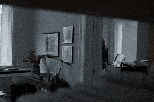 Lydia, wearing a collared shirt and sweater, walks into a room and heads for the closet, past a longhaired woman standing just offscreen, beside the piano.