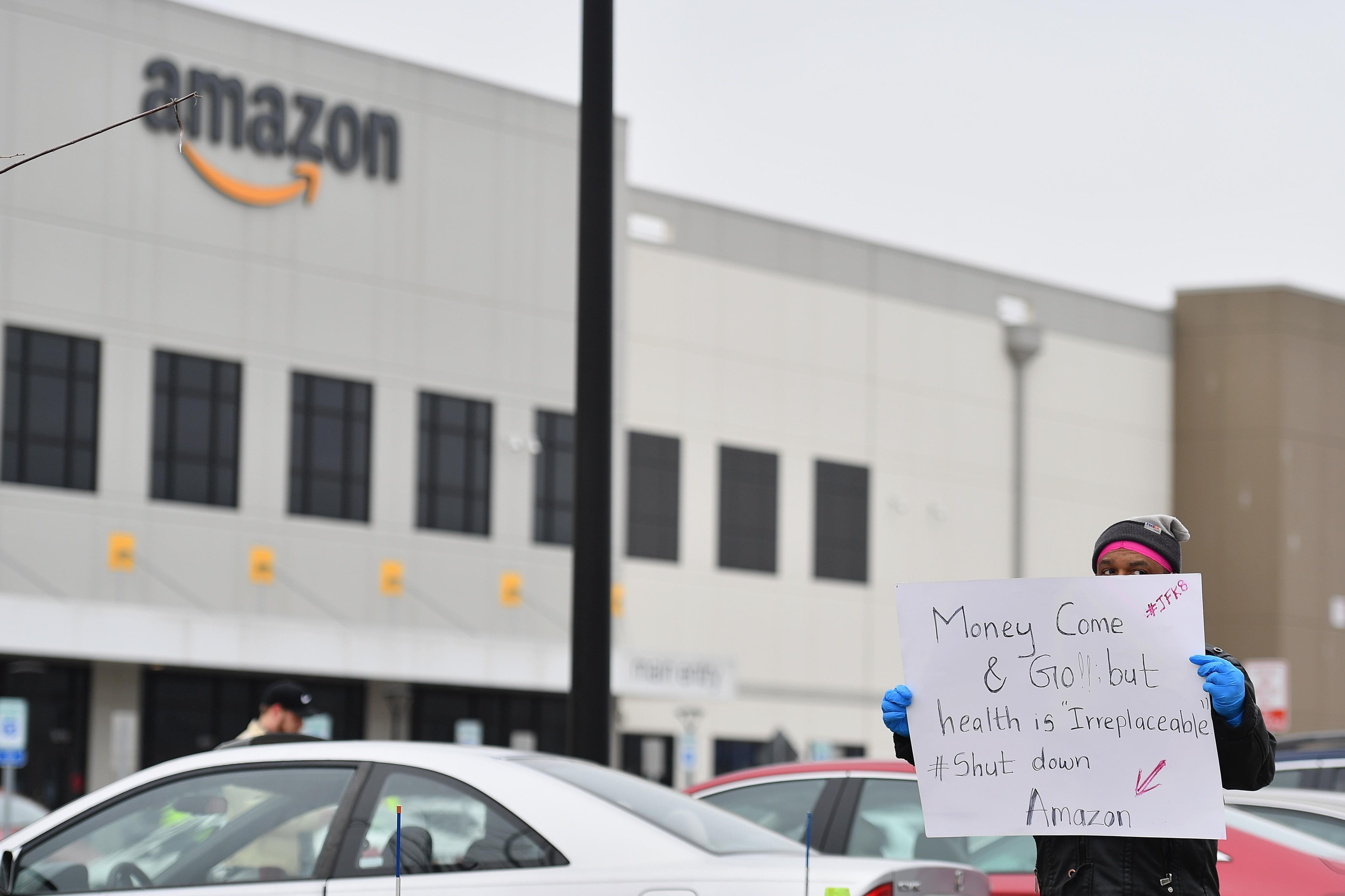 A protester stands in front of an Amazon facility holding a sign that reads "Money come and go but health is irreplaceable #Shut down Amazon"
