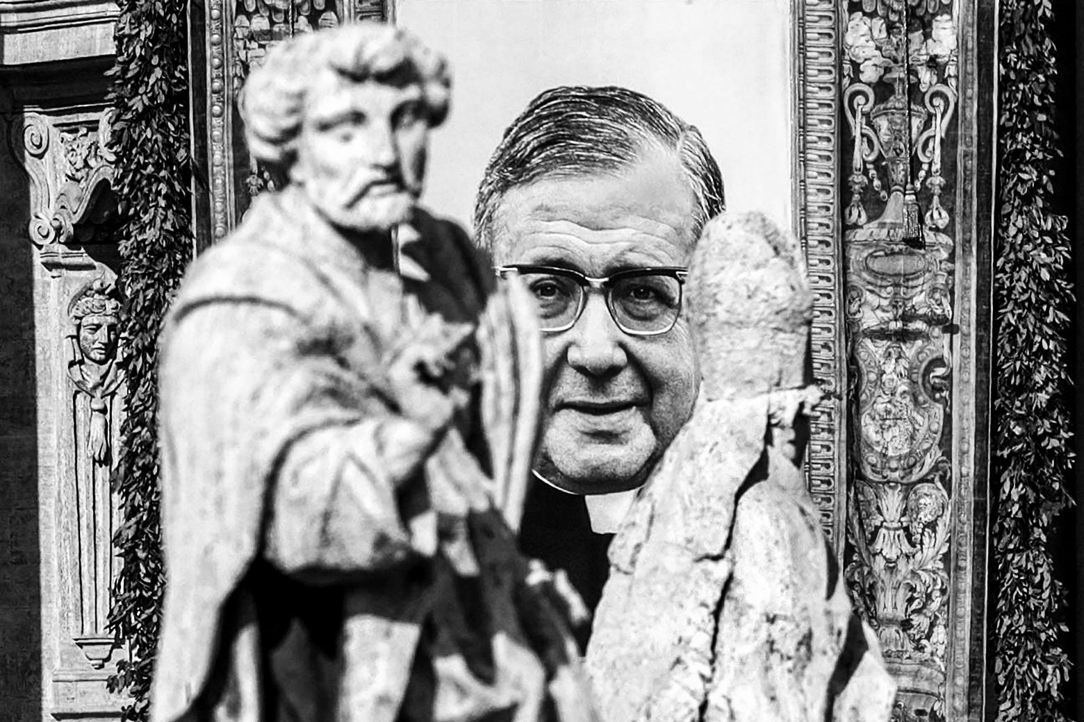 A man in glasses and a priest’s collar seen in a picture on a wall behind two statues.