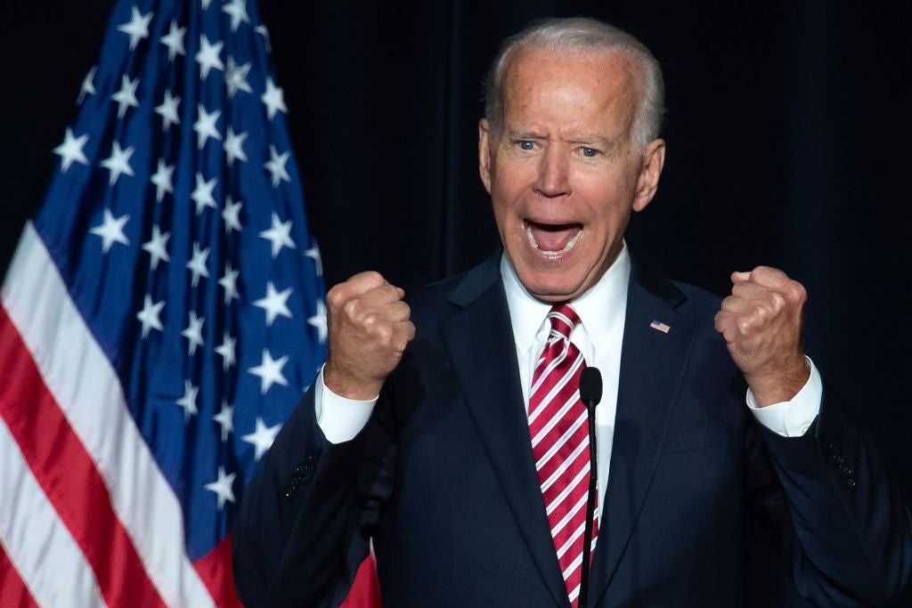 Biden clenches his fists while wearing a suit and tie and standing at a lectern in front of an American flag.