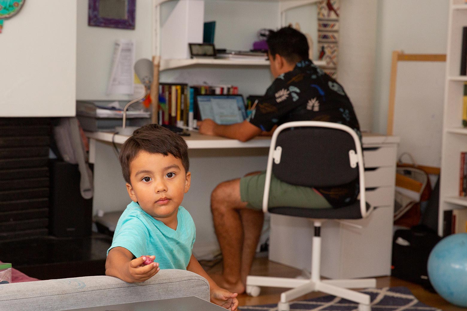 A boy looks toward the camera, reaching forward with a toy in his hand. Behind him, Oscar works on a laptop at a desk.