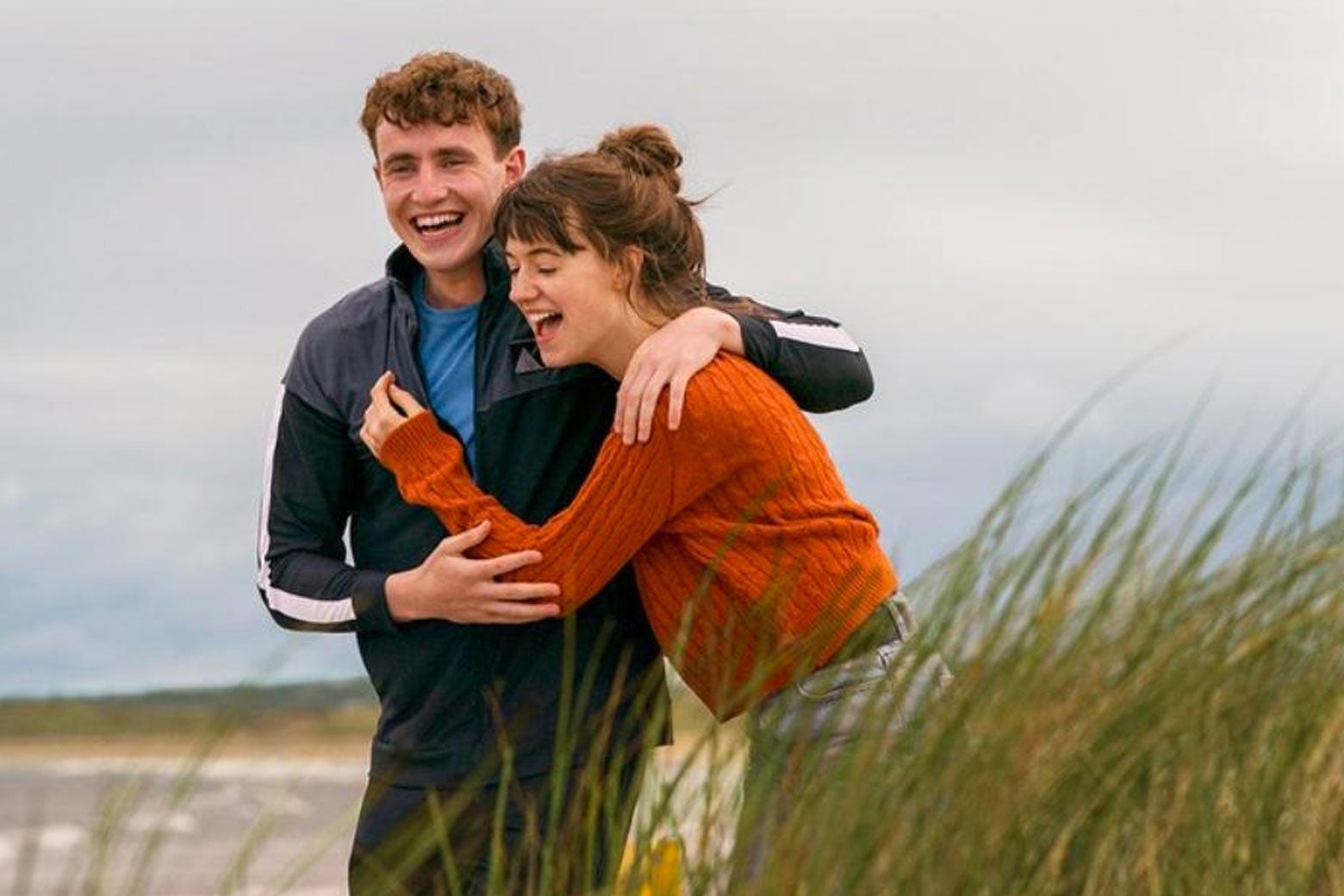 Paul Mescal and Daisy Edgar-Jones walk along a windy beach while embracing.