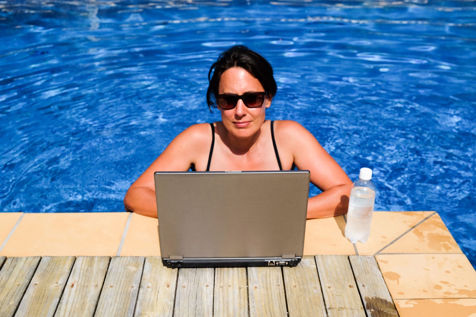 A woman in a swimming pool uses a laptop on the deck of the pool.