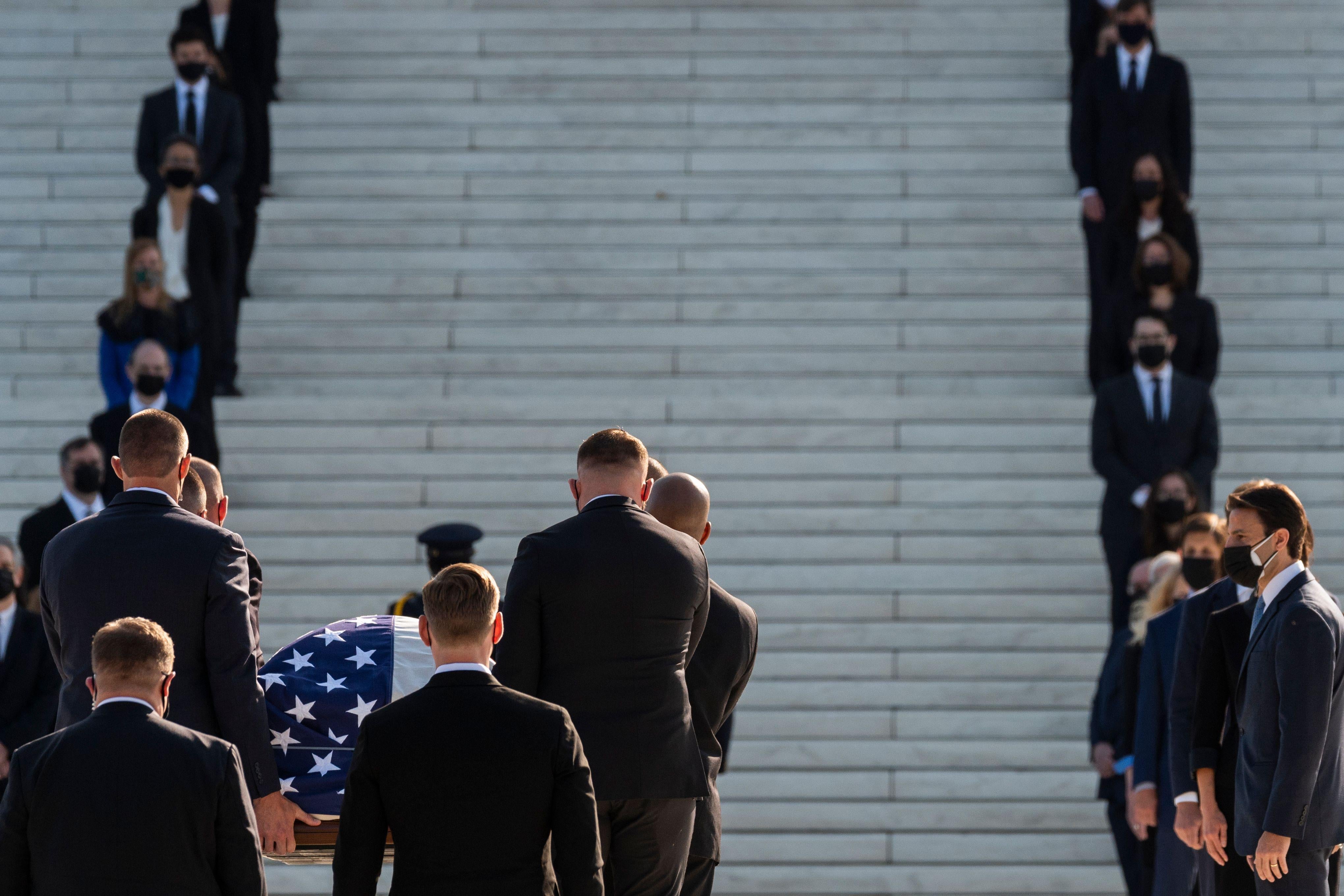 People in suits stand on the steps of the Supreme Court as a casket with an American flag is carried up the steps.
