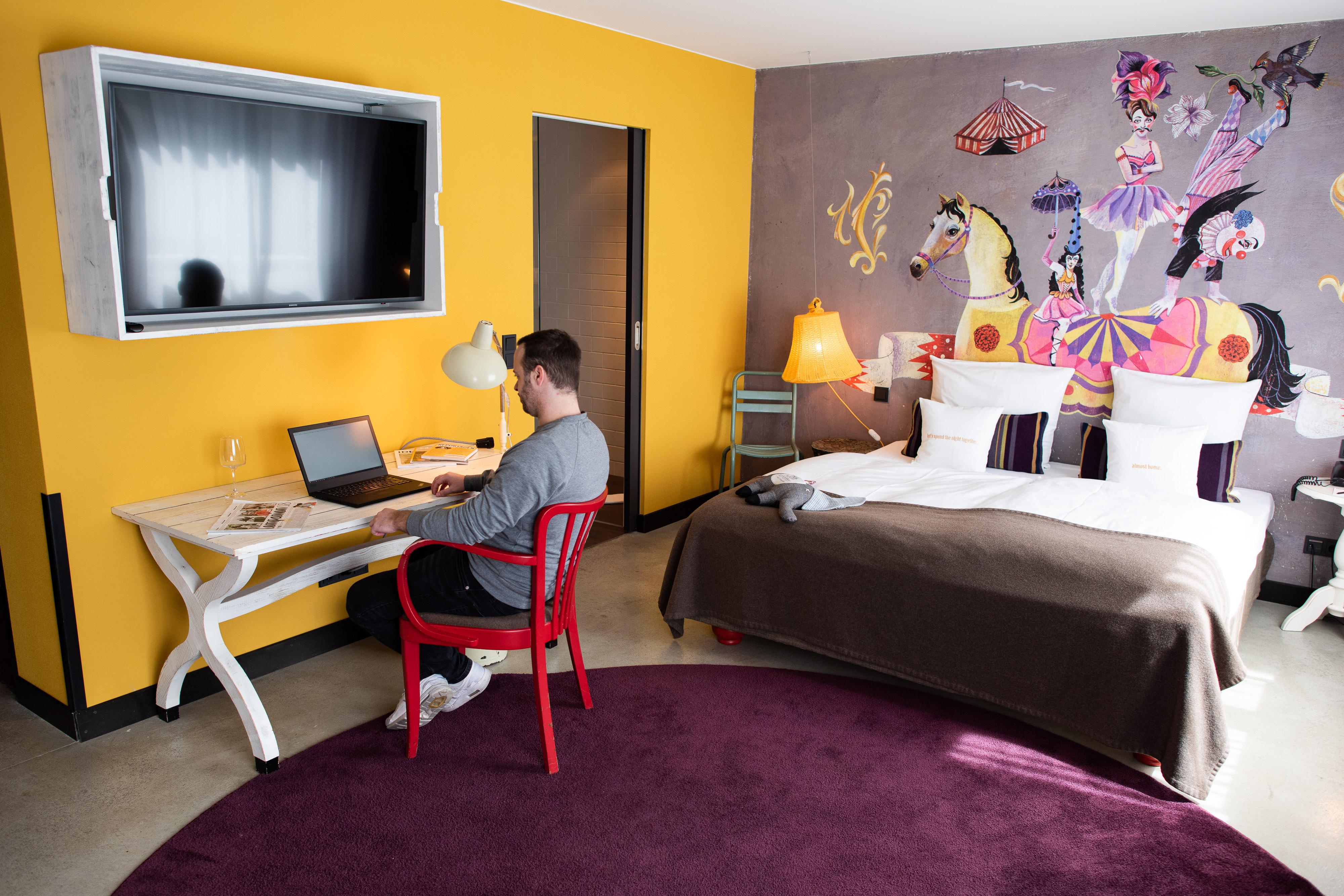 A man sits at a desk, typing at a laptop, underneath a TV screen. Behind him, a huge bed sits against a well decorated wall.