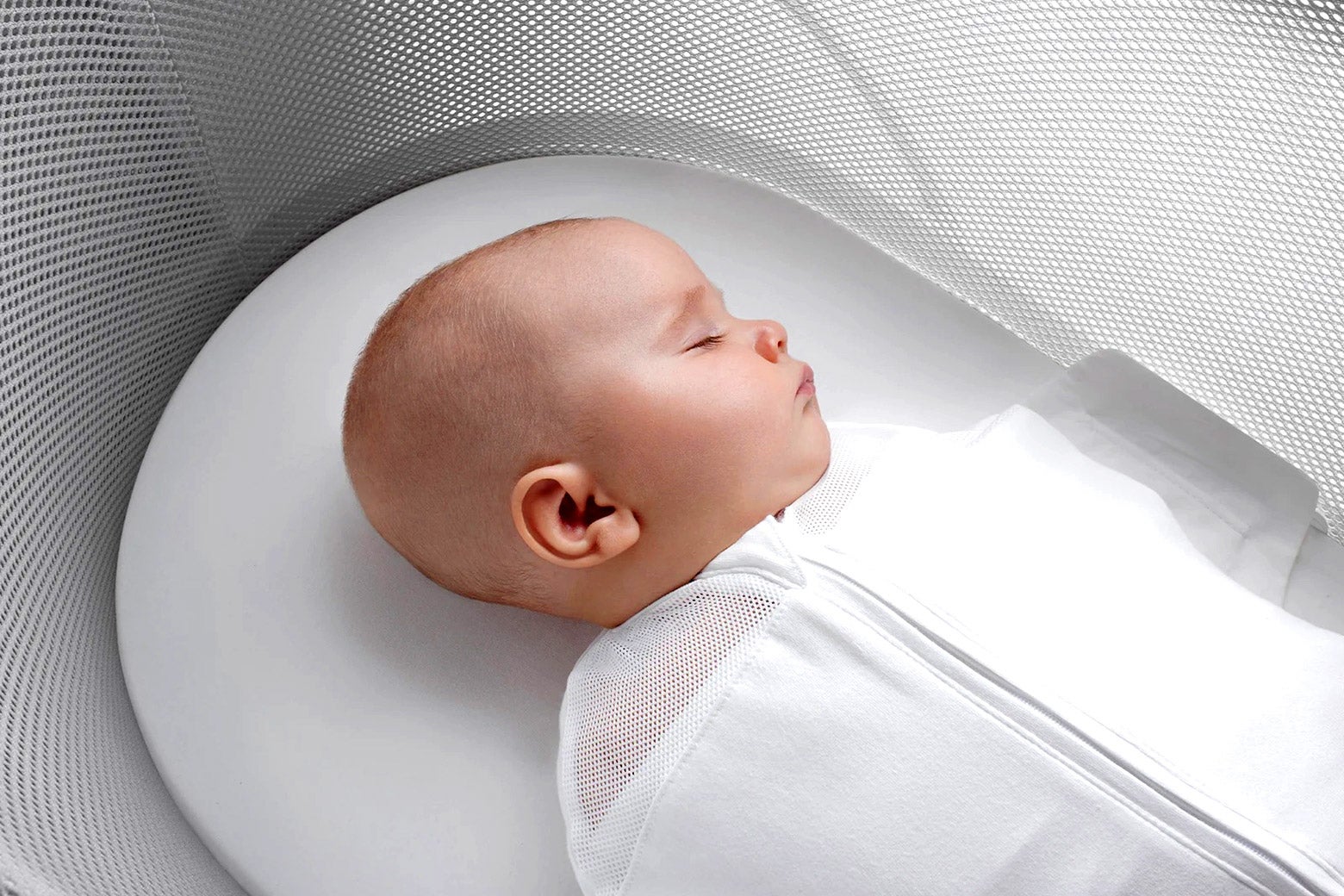A white baby sleeps in a white bassinet. It is strapped in on its back via some mesh fabric. 
