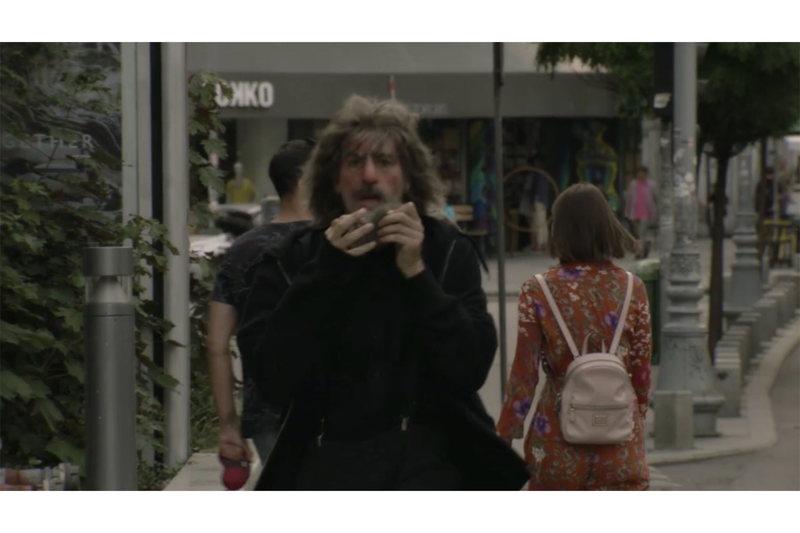 A still from Borat showing Borat walking toward camera on a city street, in front of a Yokko shop.