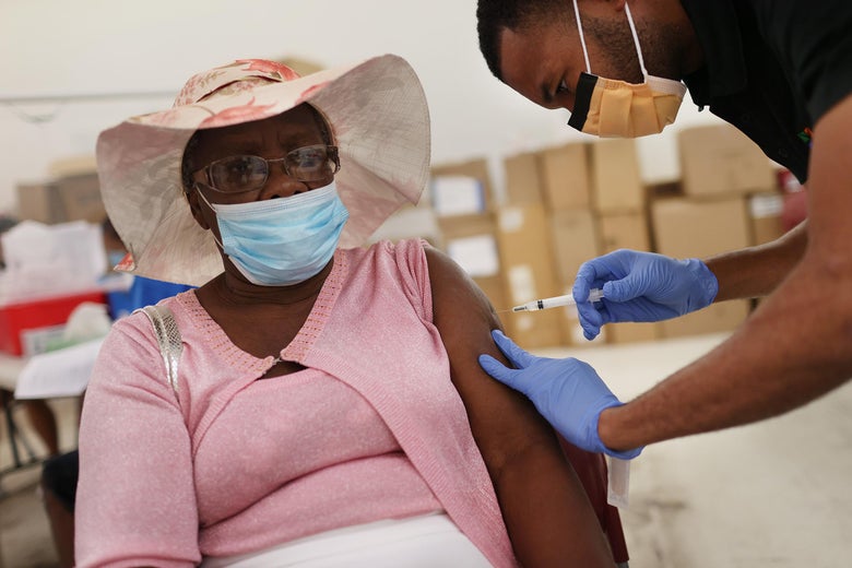 A Black man wearing gloves and a surgical mask bends over to put a shot into the arm of a seated older Black woman wearing a hat and surgical mask