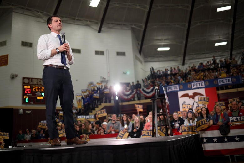 Pete Buttigieg stands on a stage holding a microphone, facing a crowd carrying signs saying his name.